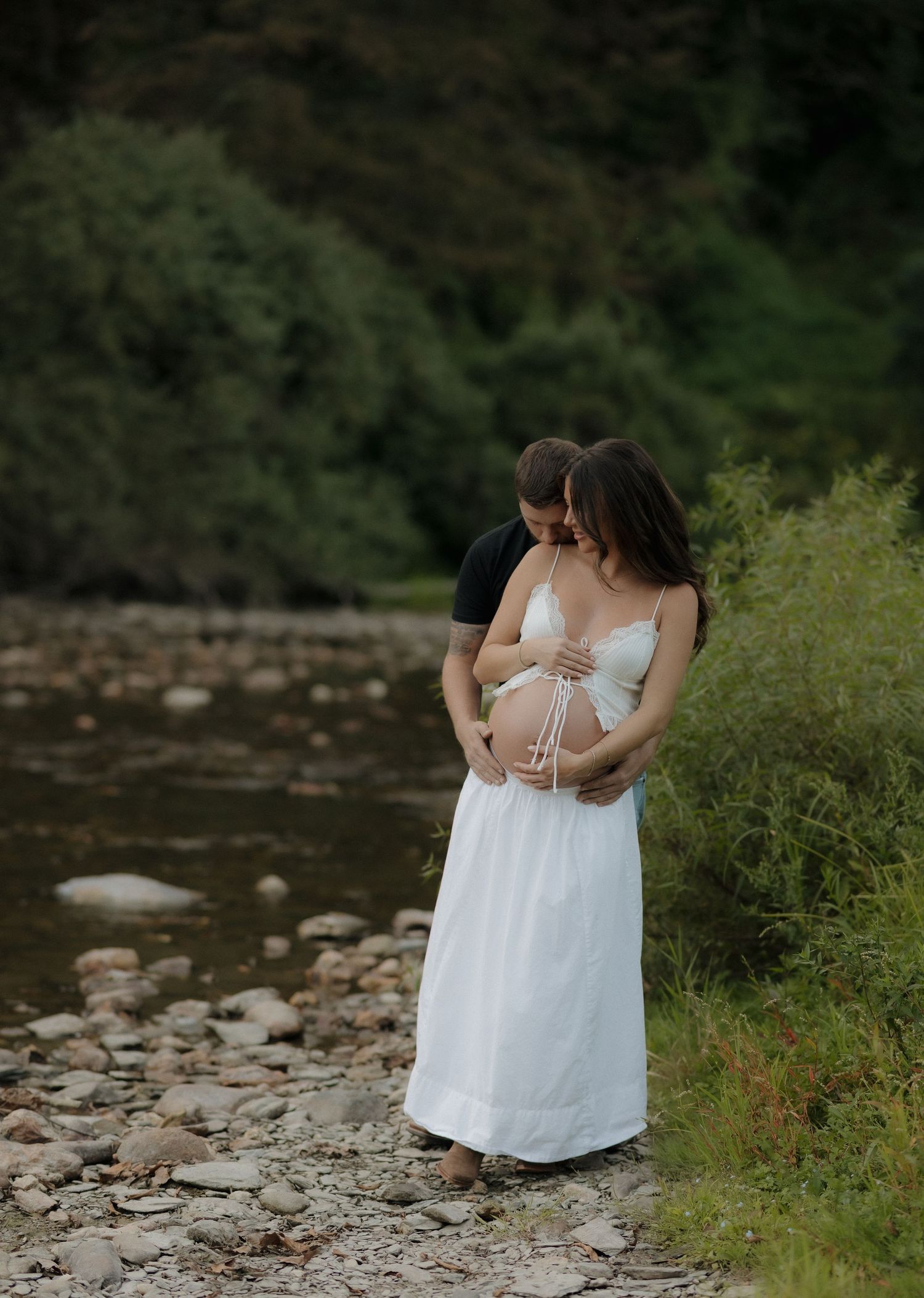 A couple shares an intimate moment during their maternity photoshoot by a rocky stream.