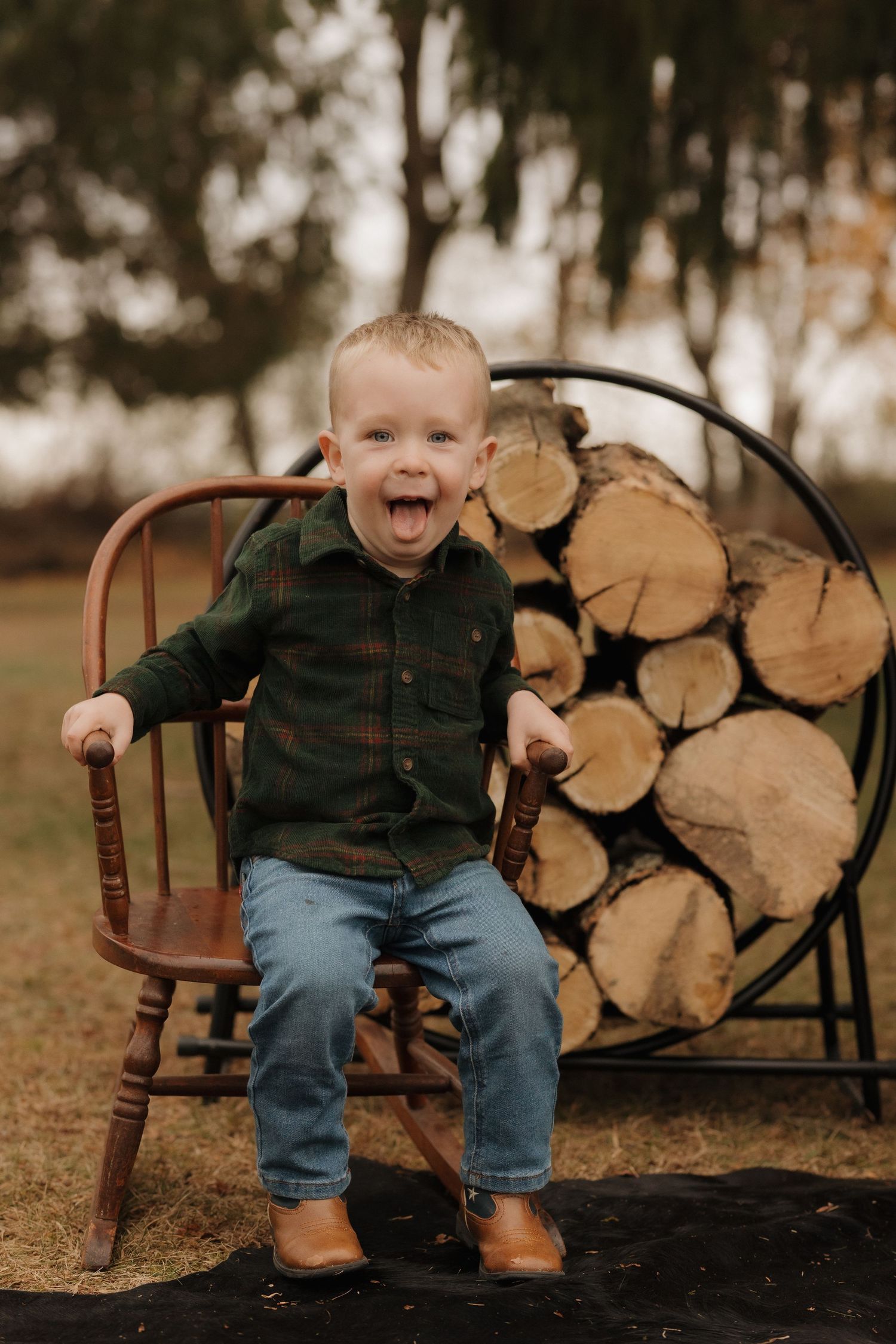 An excited toddler in green plaid sits on a wooden chair next to stacked firewood outdoors.