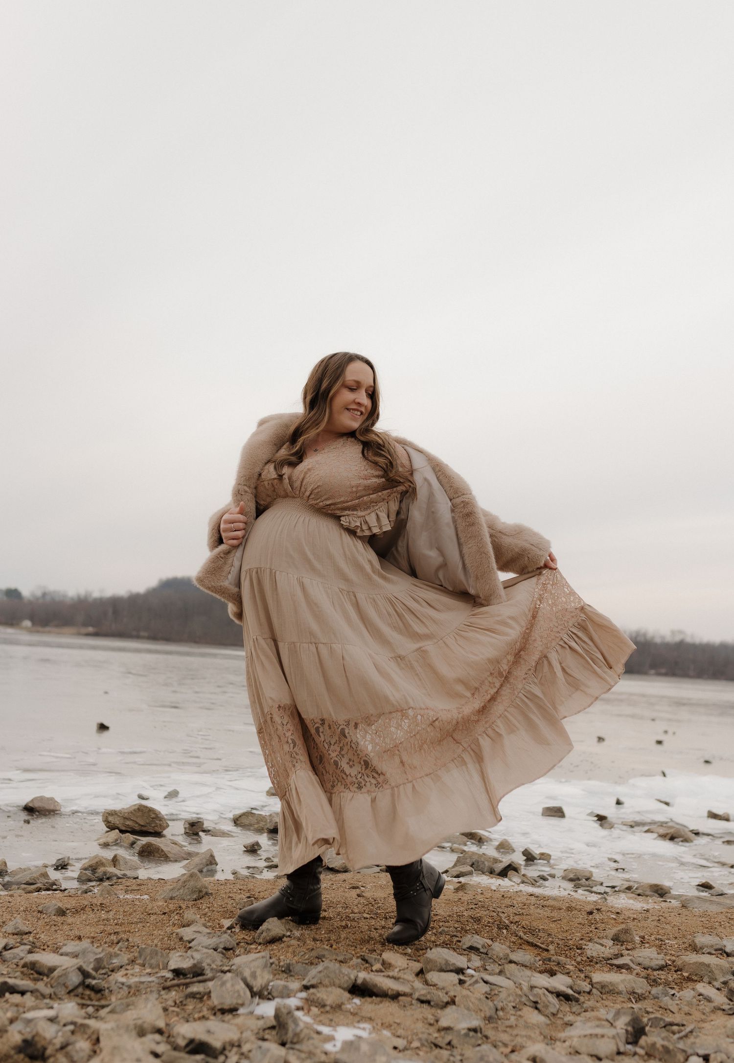 Dancing figure in beige dress twirls on rocky beach against overcast winter sky.