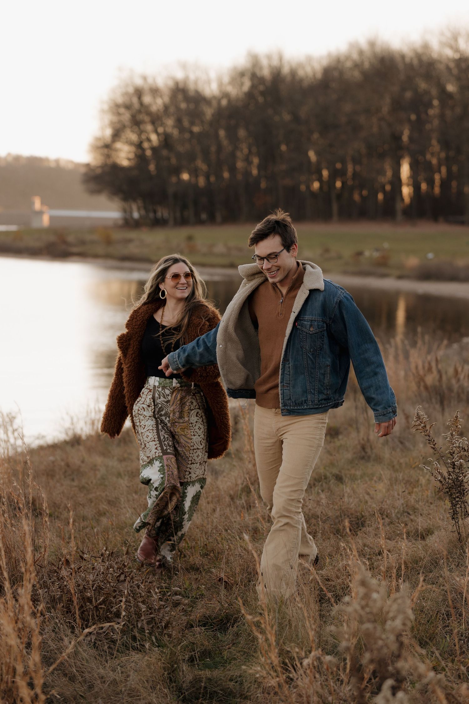 A couple walking through tall grass near a lake at golden hour.