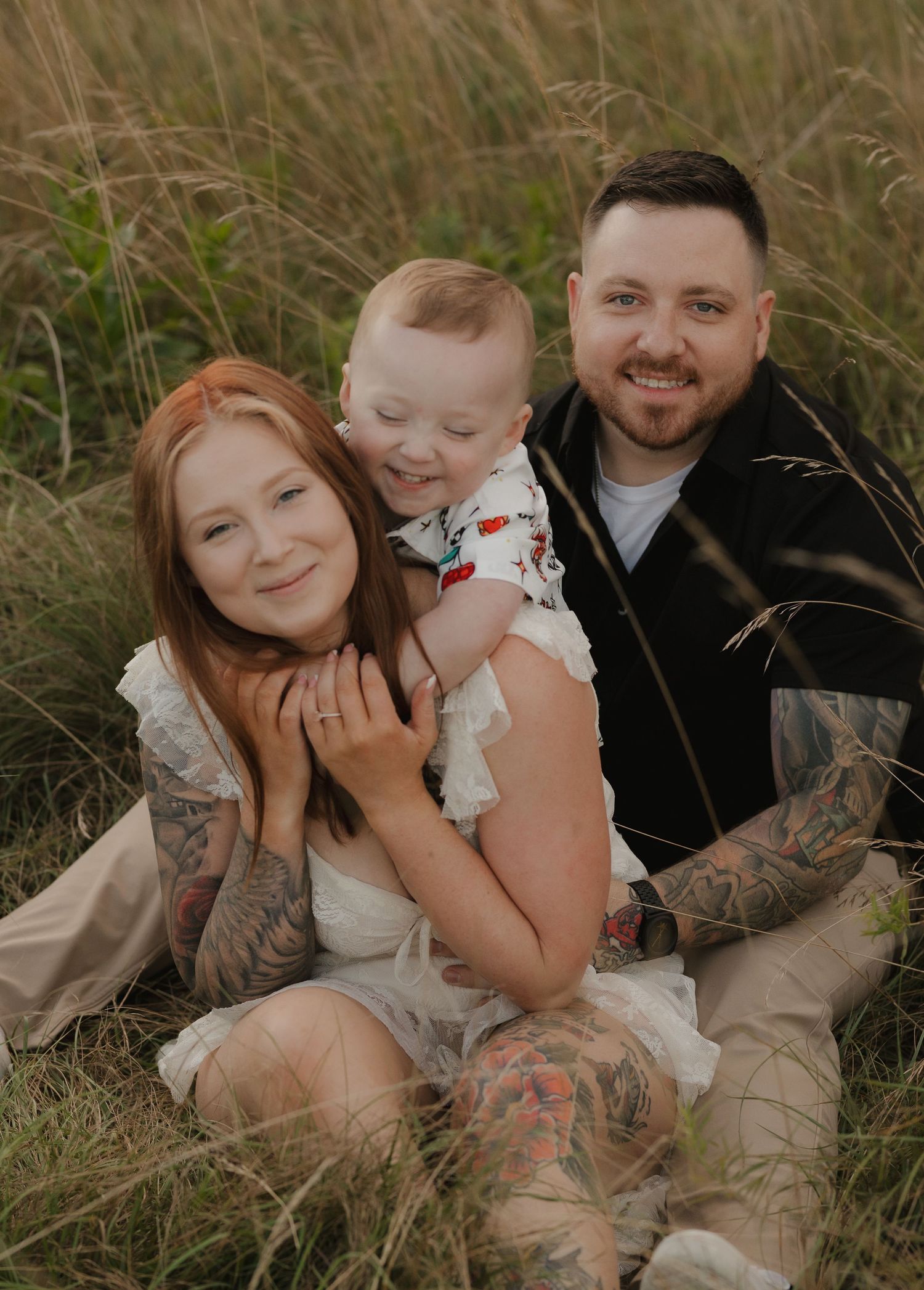 A happy family with visible tattoos poses together in tall grass during an outdoor photoshoot.