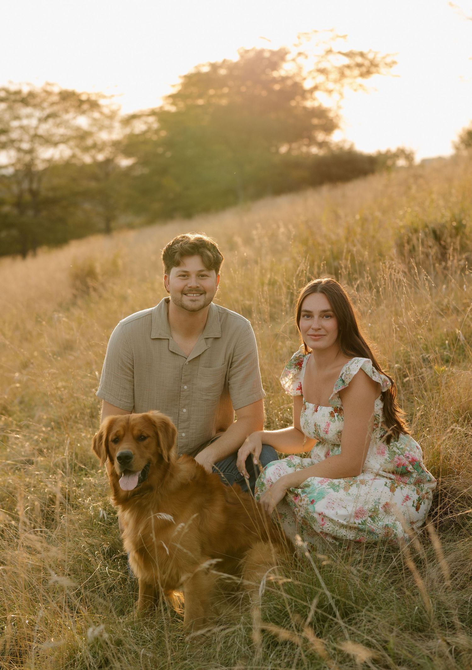 Couple and their golden retriever dog pose together in a sunny field at sunset.