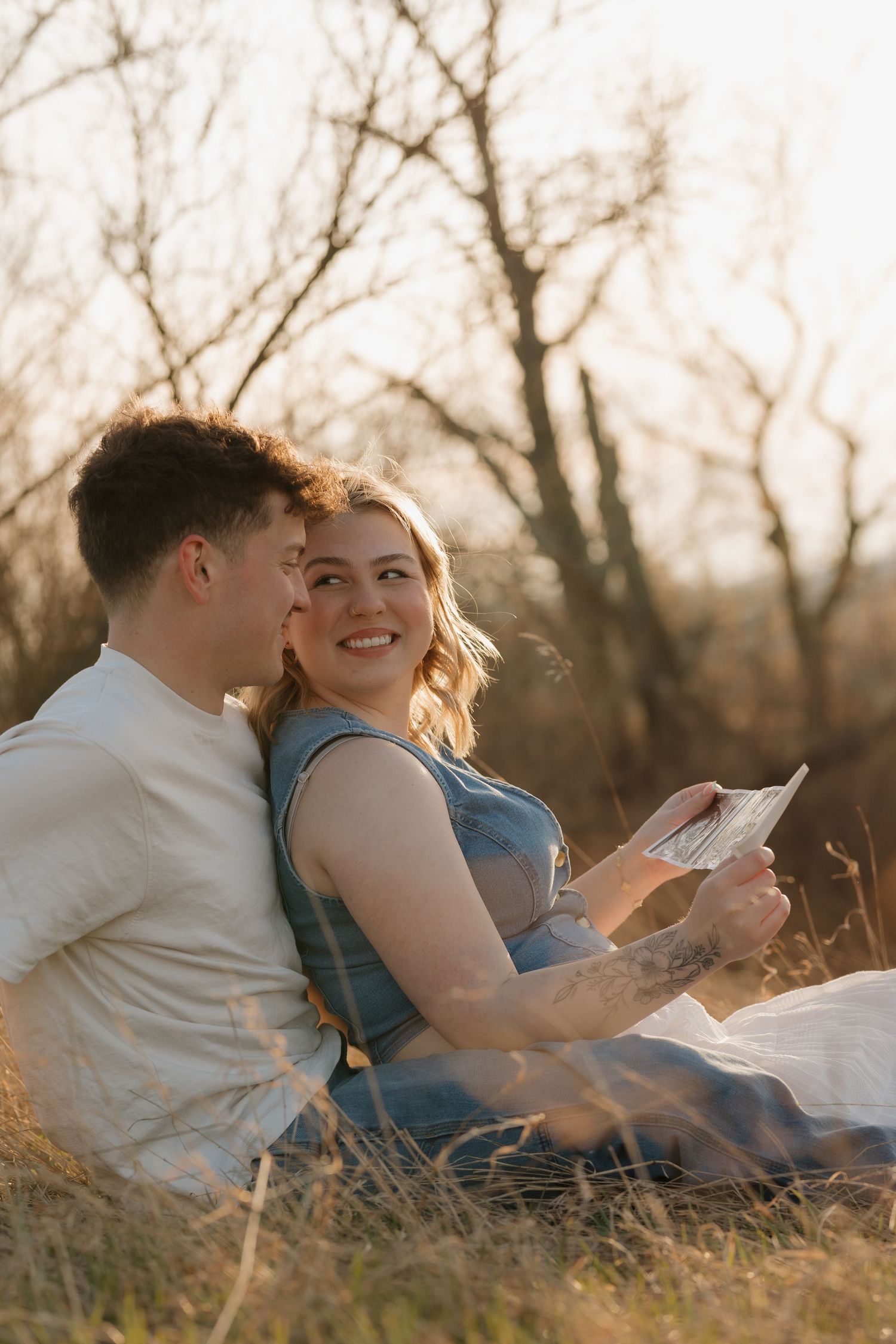 A couple sits together in a sunlit field reading a book during golden hour as trees create a dreamy backdrop.