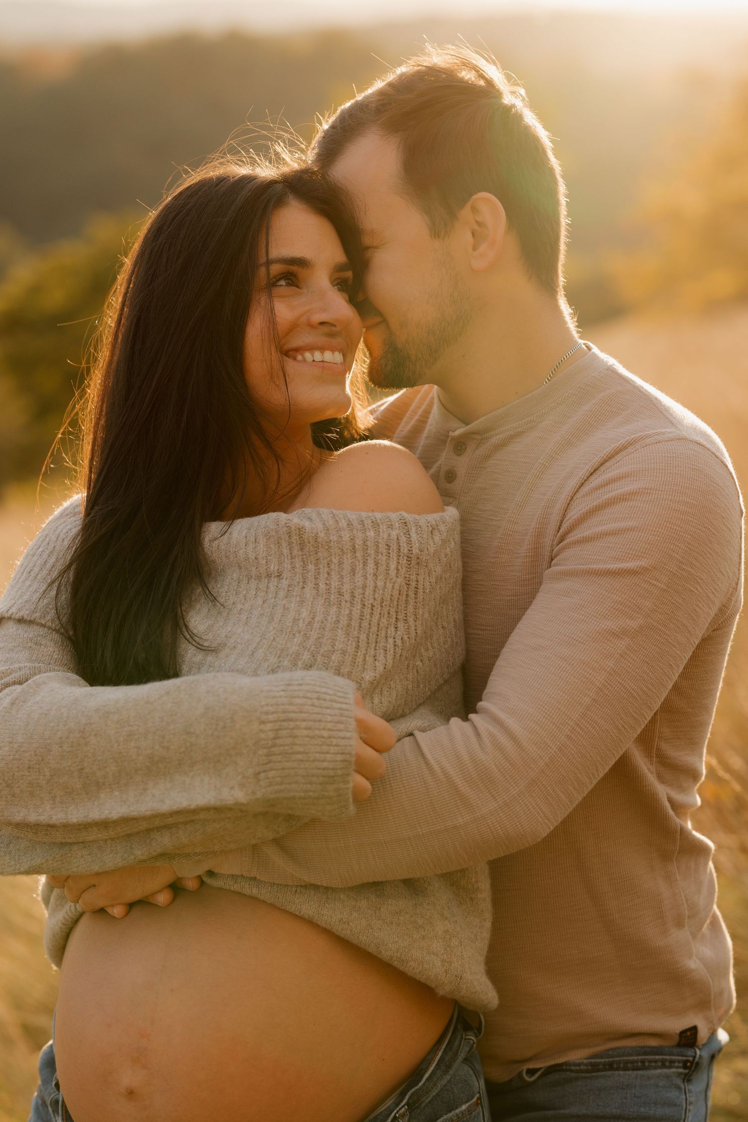 Couple in cozy sweaters embracing during golden hour sunset photoshoot. Maternity