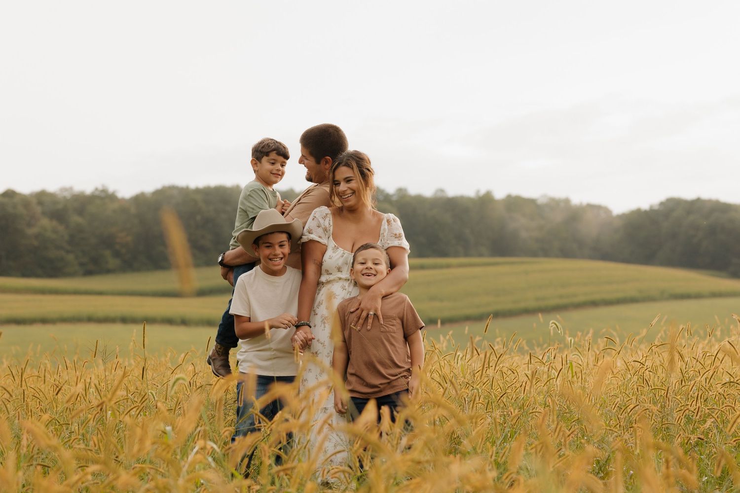 Family of five smiling together in a golden wheat field with lush green trees in the background.
