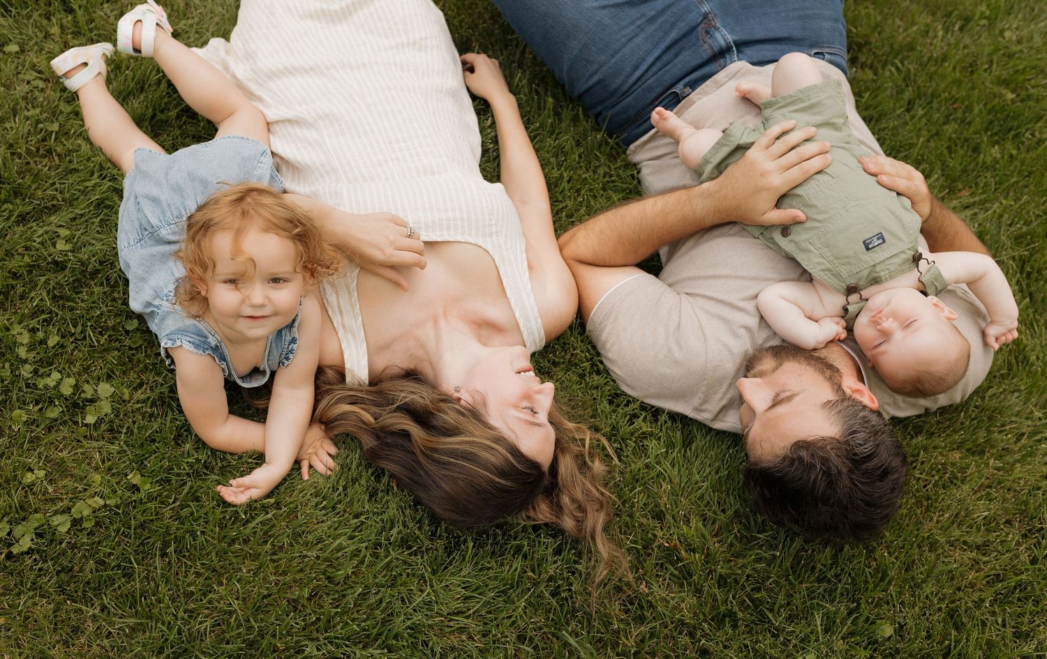 Family of four lying on green grass, parents with toddler girl and baby enjoying a sunny outdoor moment.