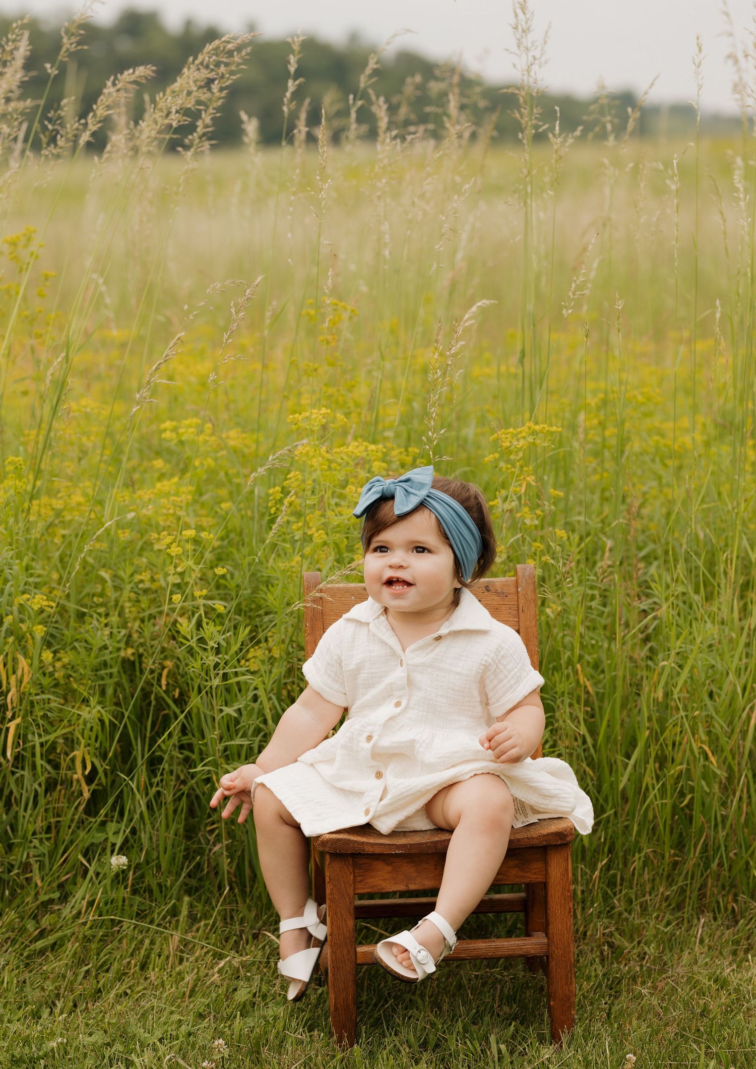 Baby in white dress and blue headband sitting on wooden bench in tall grass field.