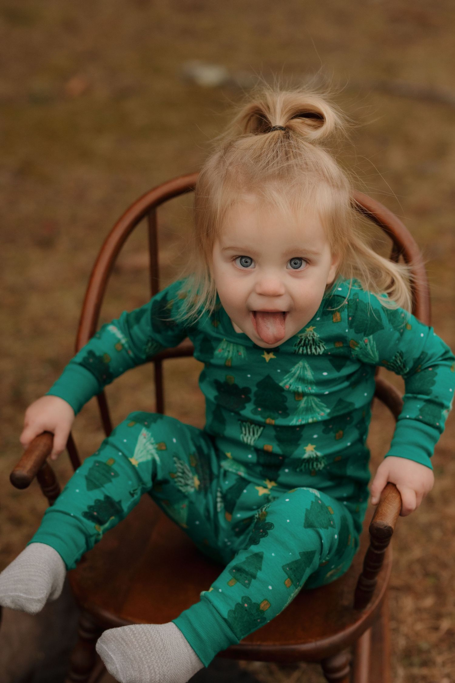A toddler in green dinosaur pajamas makes a silly face while sitting in a wooden chair.