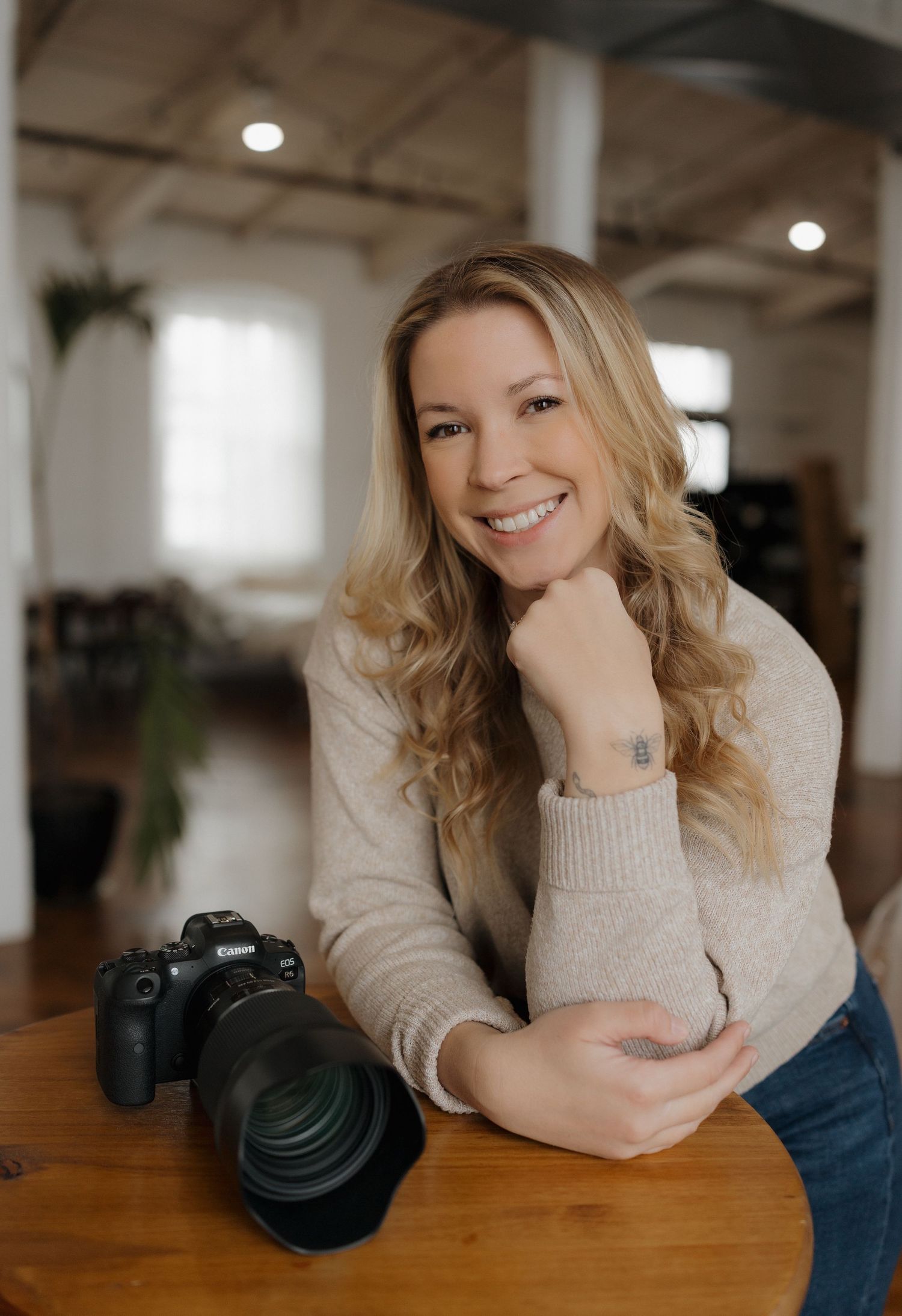 Professional photographer in beige sweater leaning on wooden table with DSLR camera and telephoto lens.