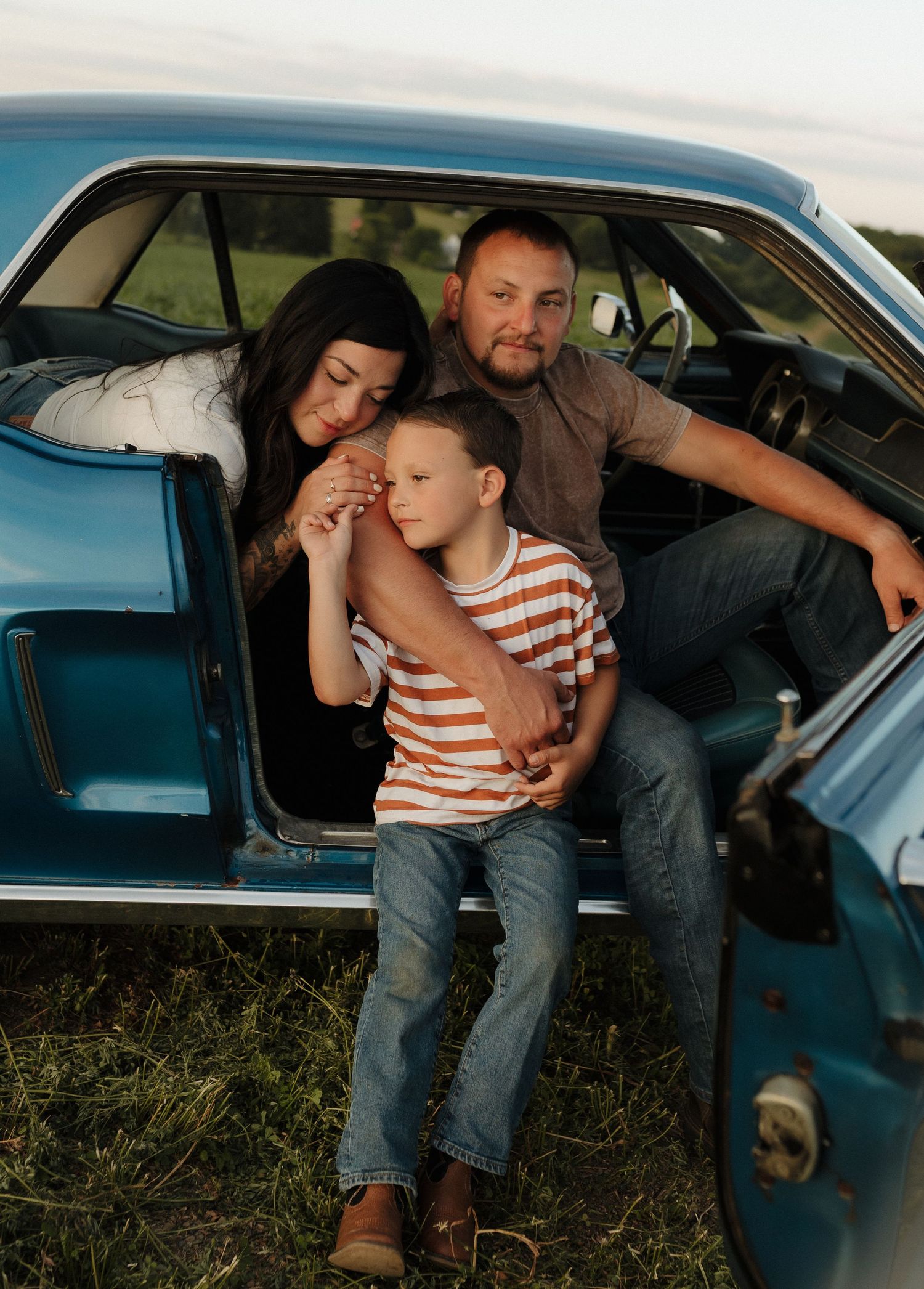 Family of three posing in a vintage blue car outdoors, parents embracing young boy during a golden hour photo session.