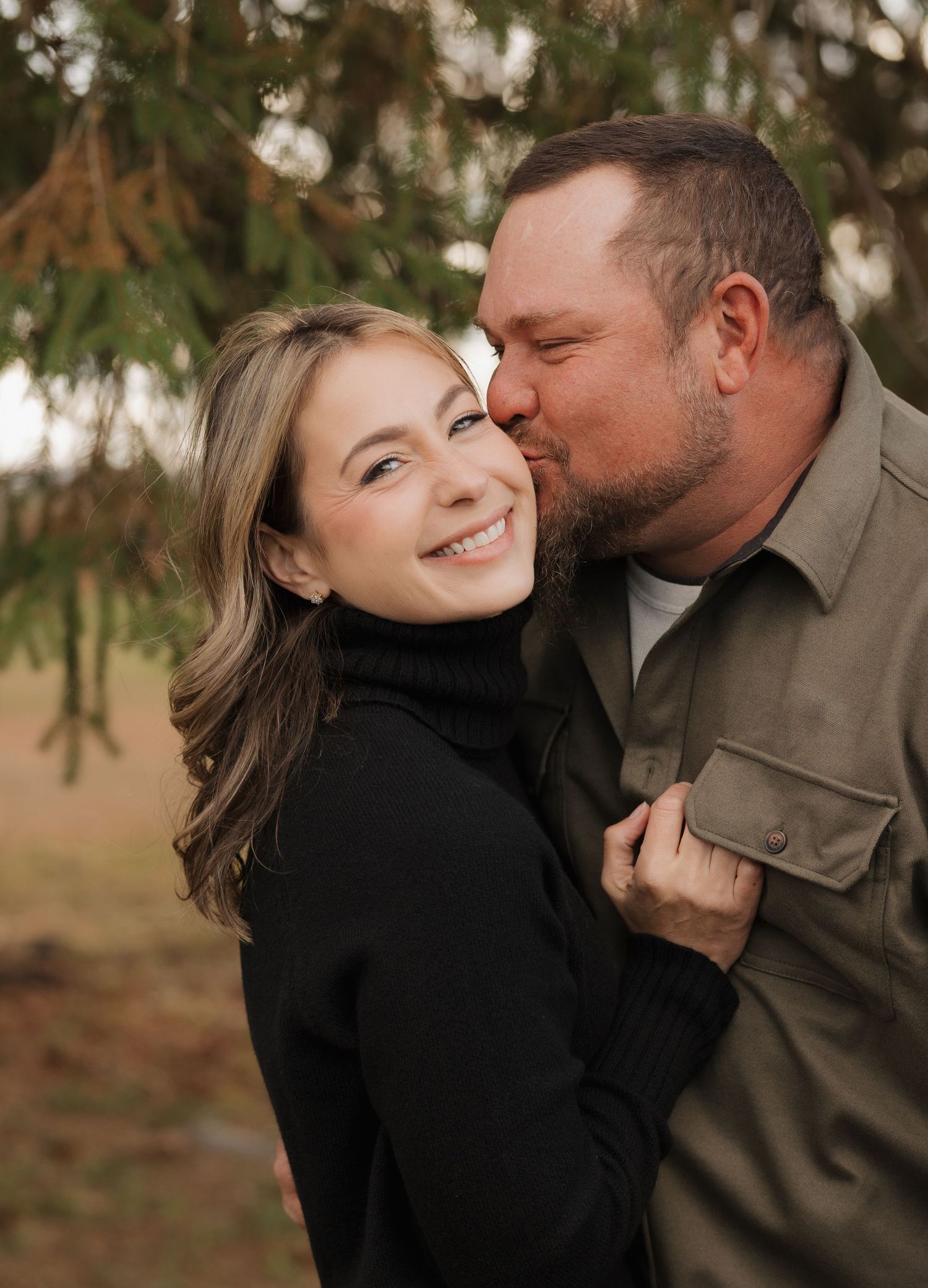 A romantic moment captured between a couple in casual attire against an evergreen backdrop.