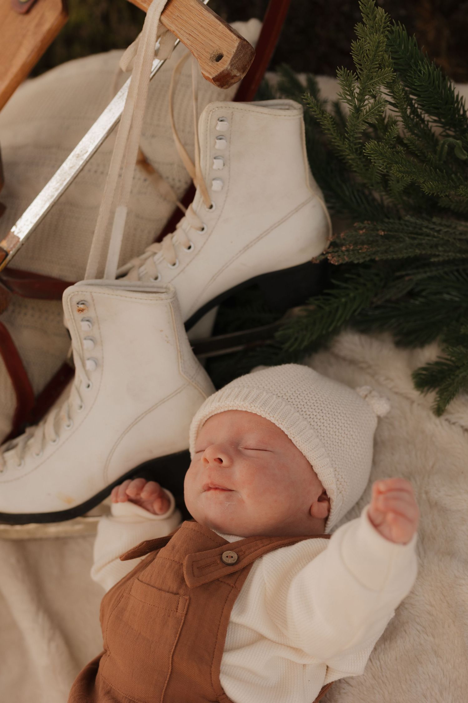 Sleeping baby in white clothing lies next to decorative white ice skates and evergreen branches.