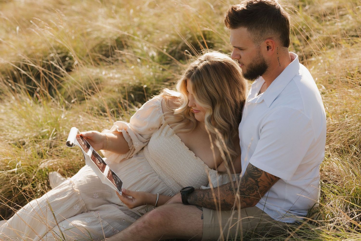 Couple sitting in golden grass field sharing intimate moments together during sunset photoshoot.