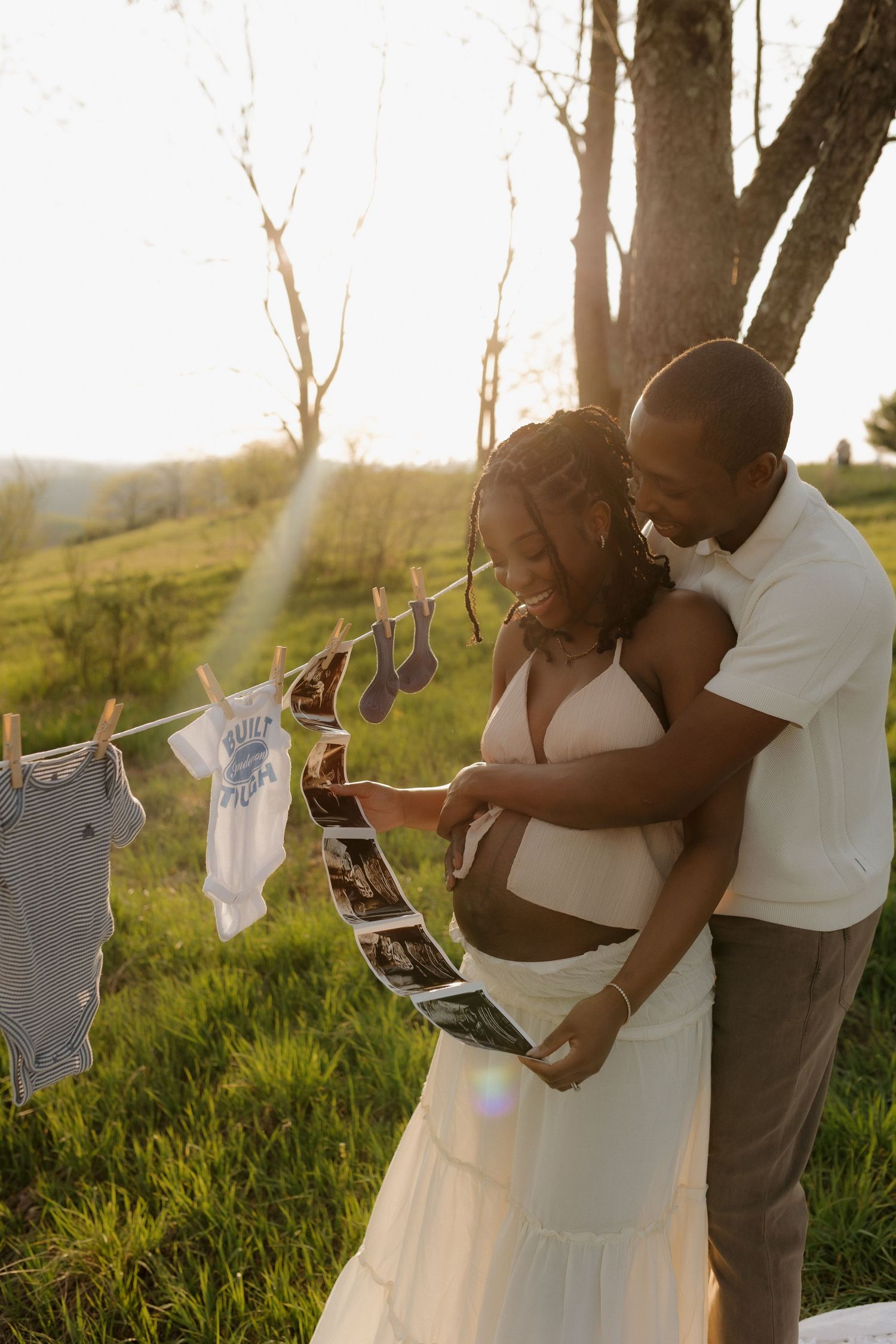 Expectant couple reviewing ultrasound photos outdoors at golden hour, with baby clothes hanging on a line nearby.