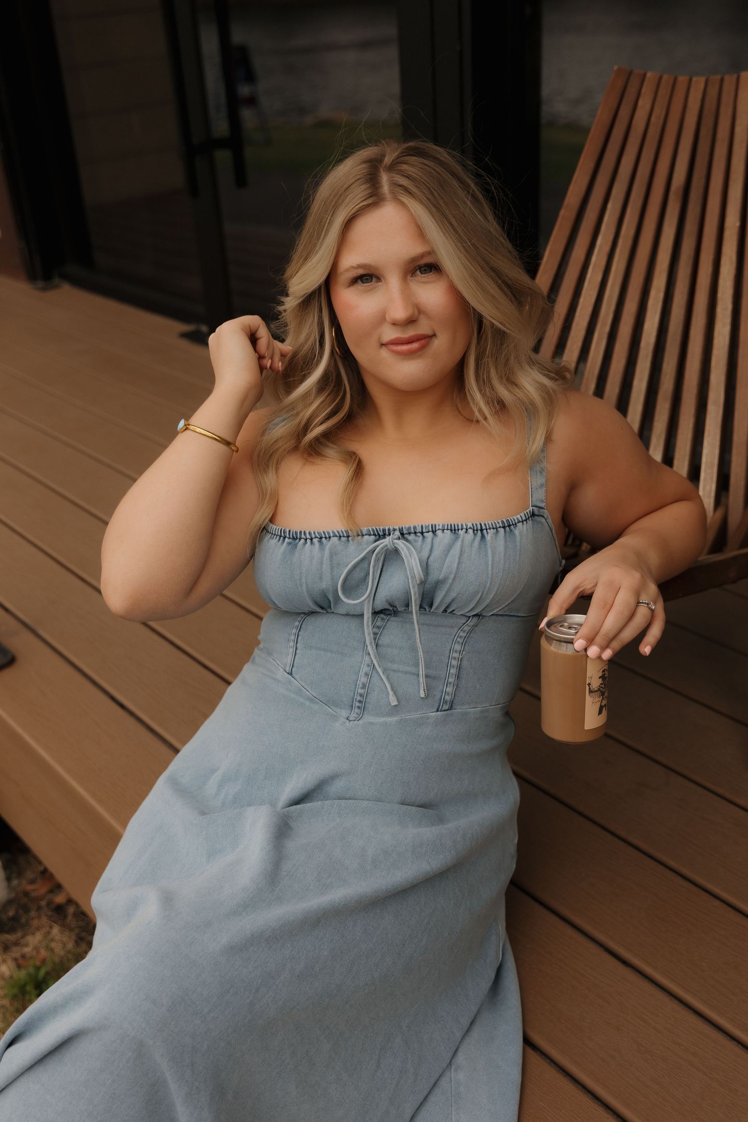 A woman in a blue dress relaxes on a wooden deck with a drink.
