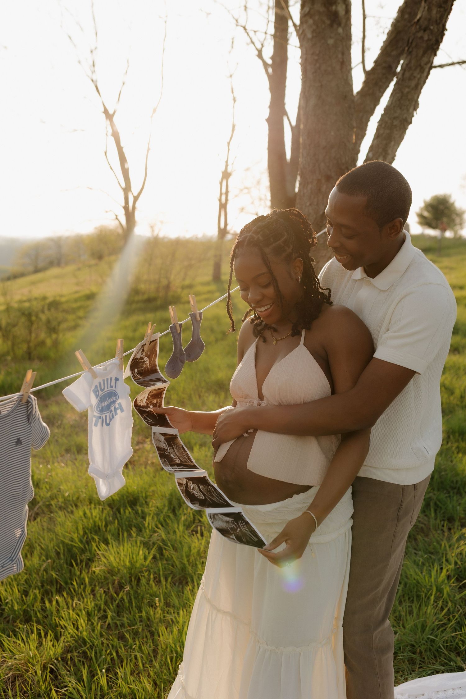A couple looks at ultrasound photos outdoors at sunset, with baby clothes hanging on a line, celebrating their pregnancy.