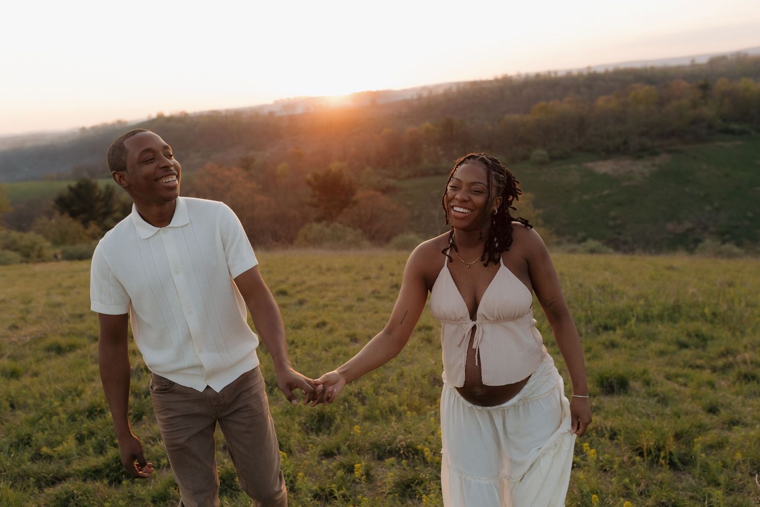 Couple holding hands walking in a green field at golden hour sunset with rolling hills in the background.