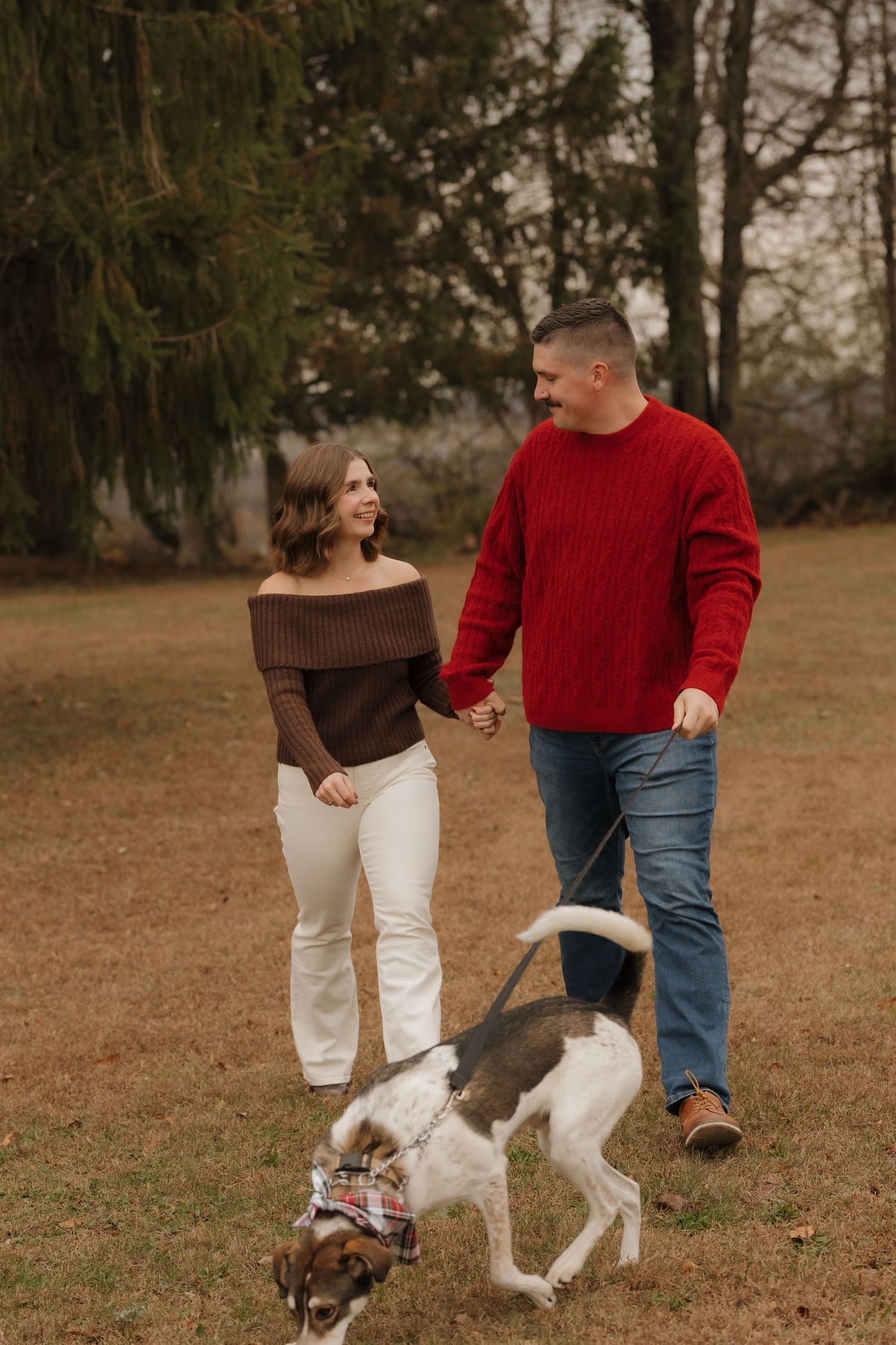 Couple in red and brown sweaters walk their dog through autumn field.