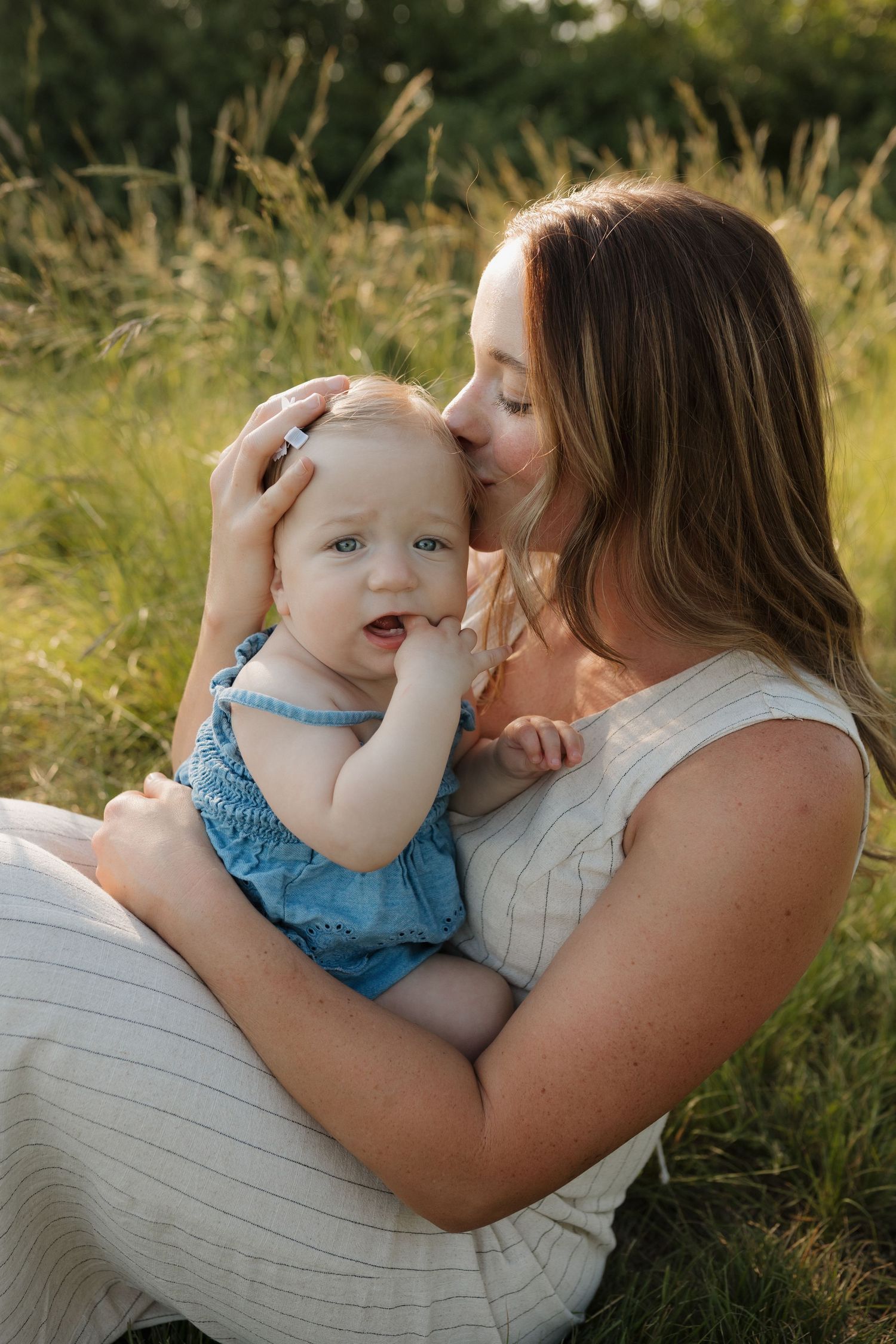 A mother tenderly kisses her smiling baby girl in a sunny green field, both dressed in light summer outfits.