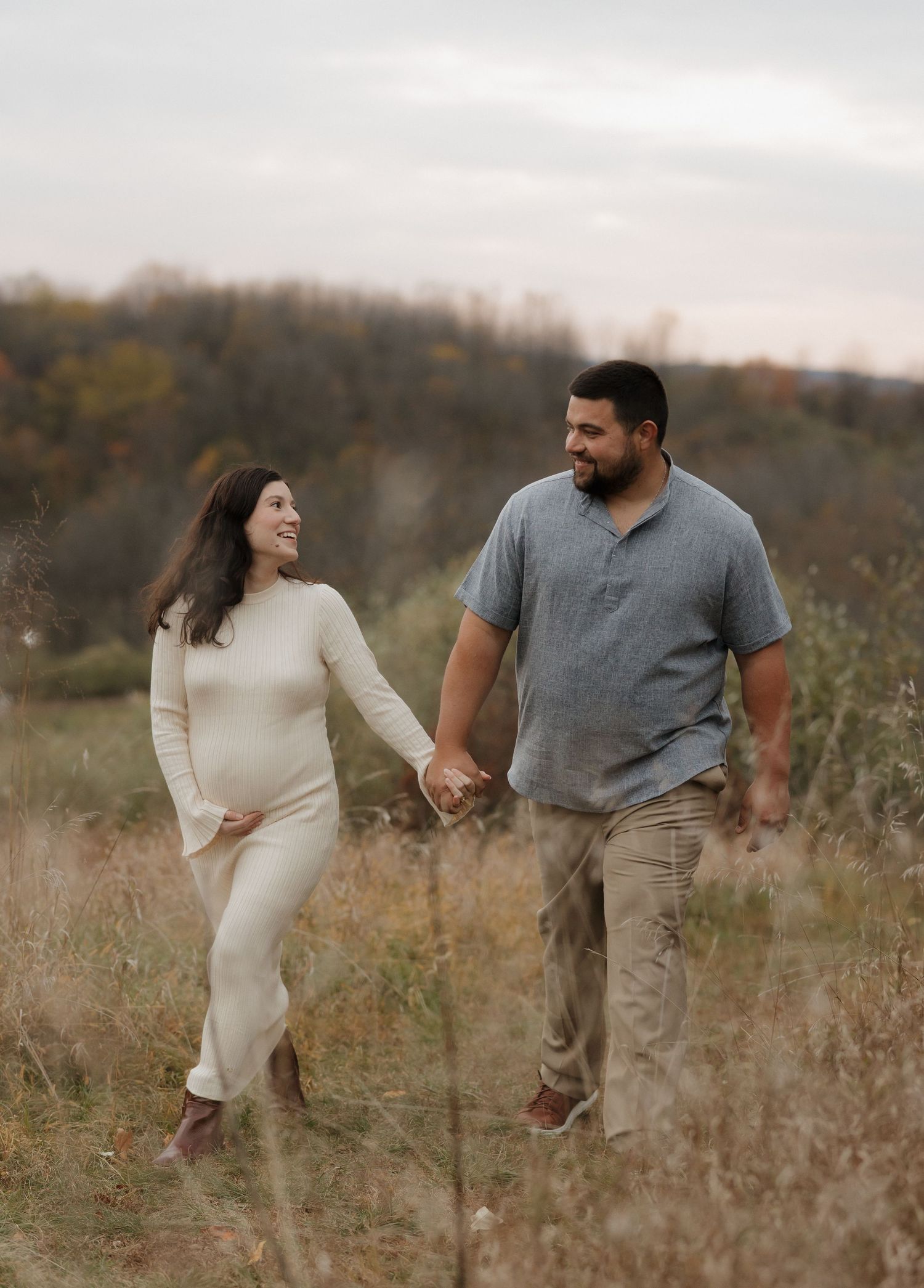 Couple walks hand in hand through tall grass in an autumn field.