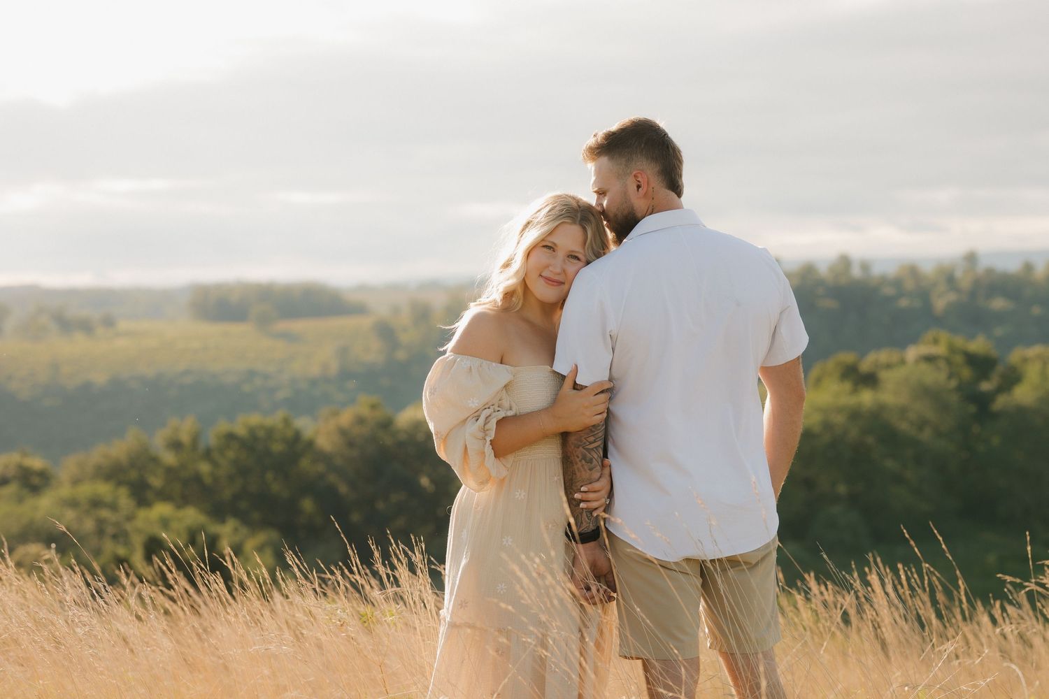 A couple embraces in a golden field during sunset while wearing casual white outfits.
