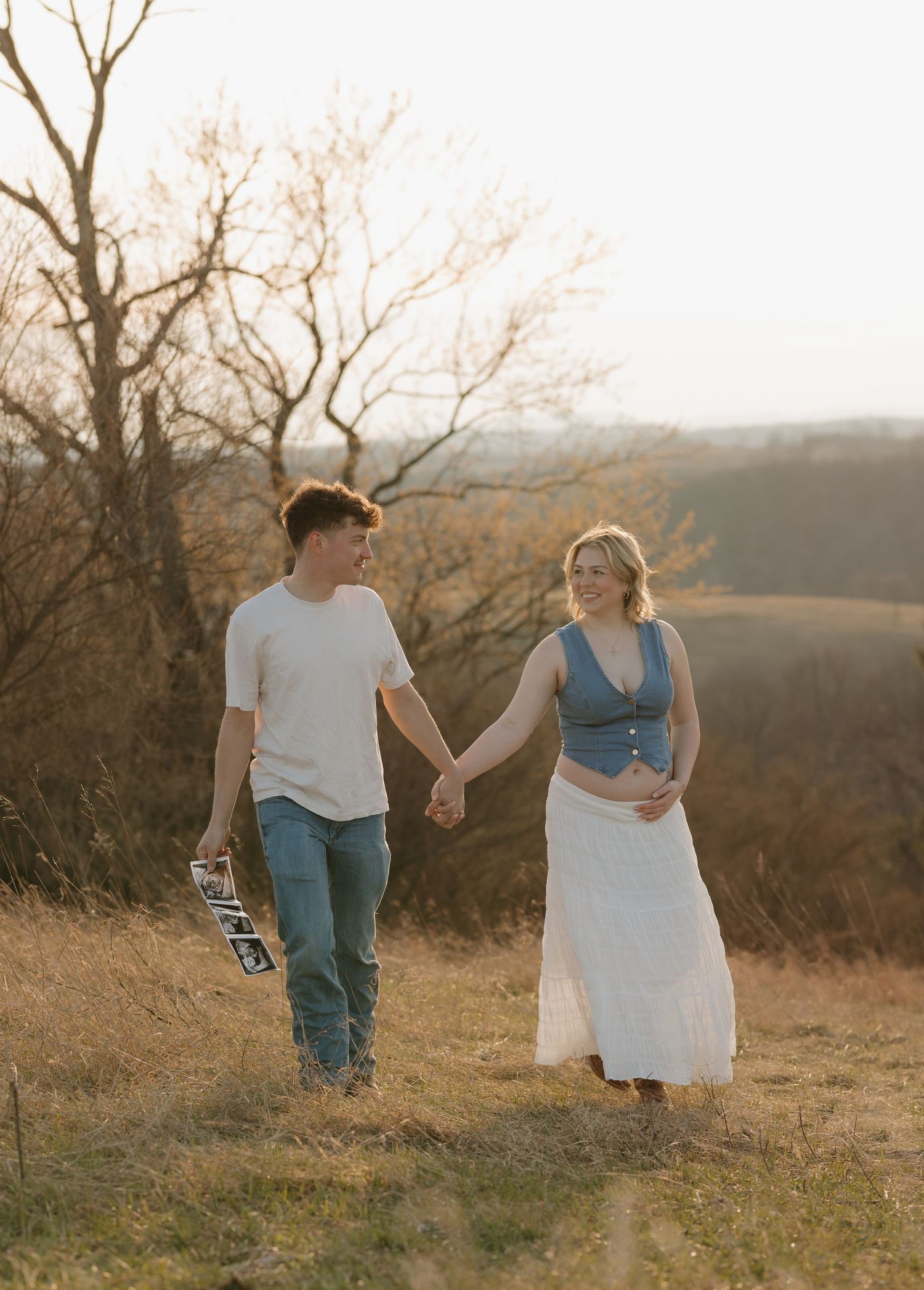 A couple walks hand in hand through a golden field at sunset during an outdoor photoshoot.