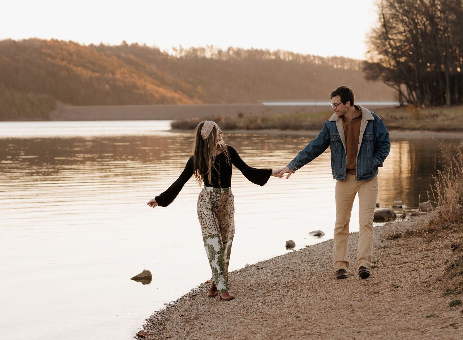 Couple walking along rocky shoreline at sunset in casual autumn outfits.