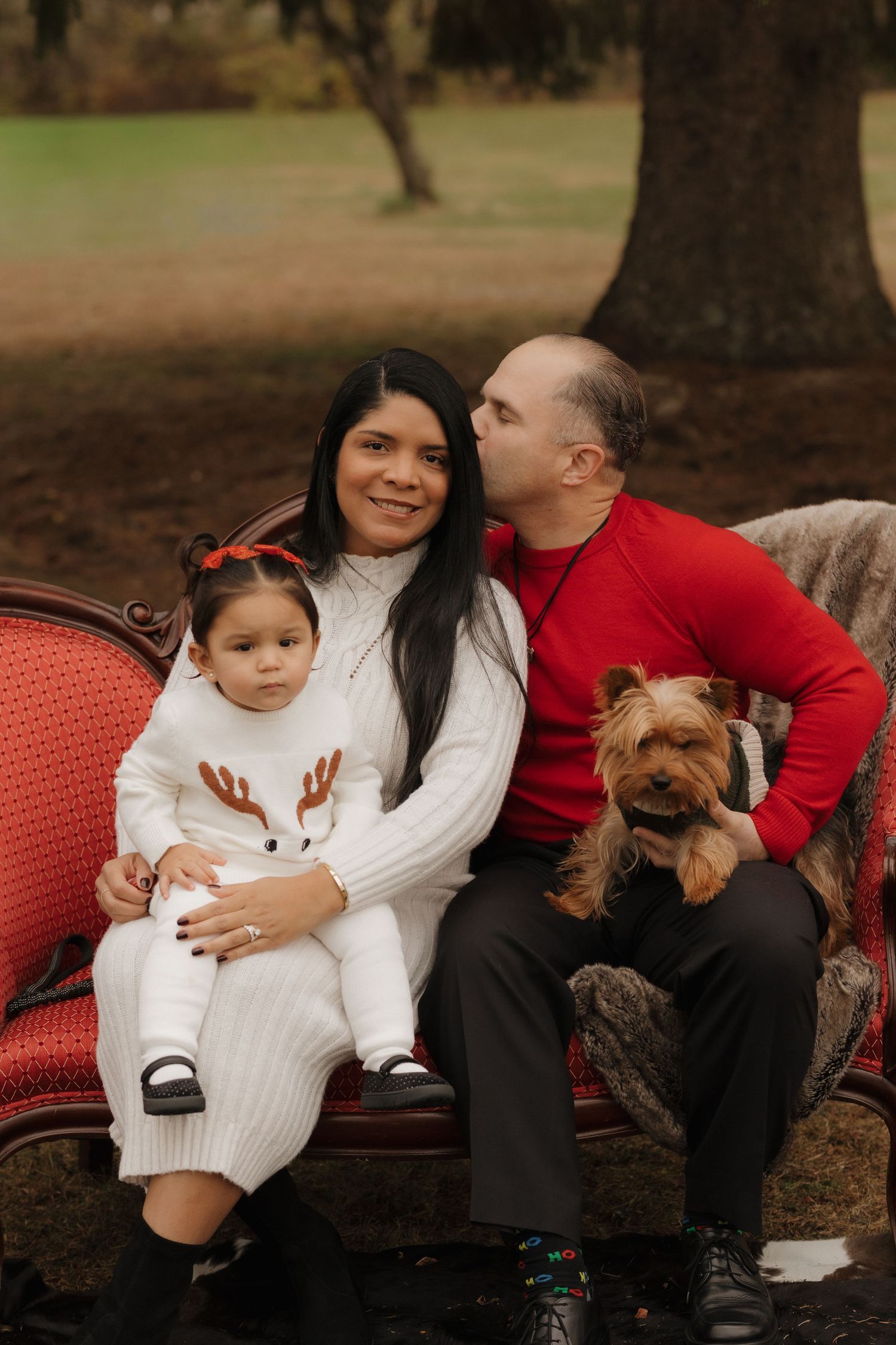 Family and their small dog relax together on a red vintage settee during outdoor photo session.