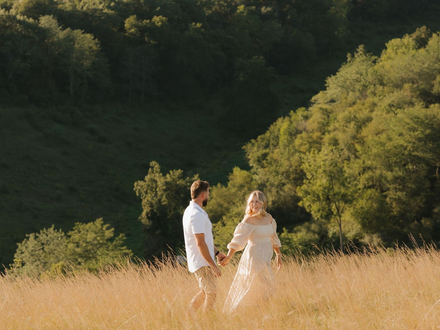 A couple in wedding attire walks through a sunlit meadow surrounded by lush green trees during golden hour.