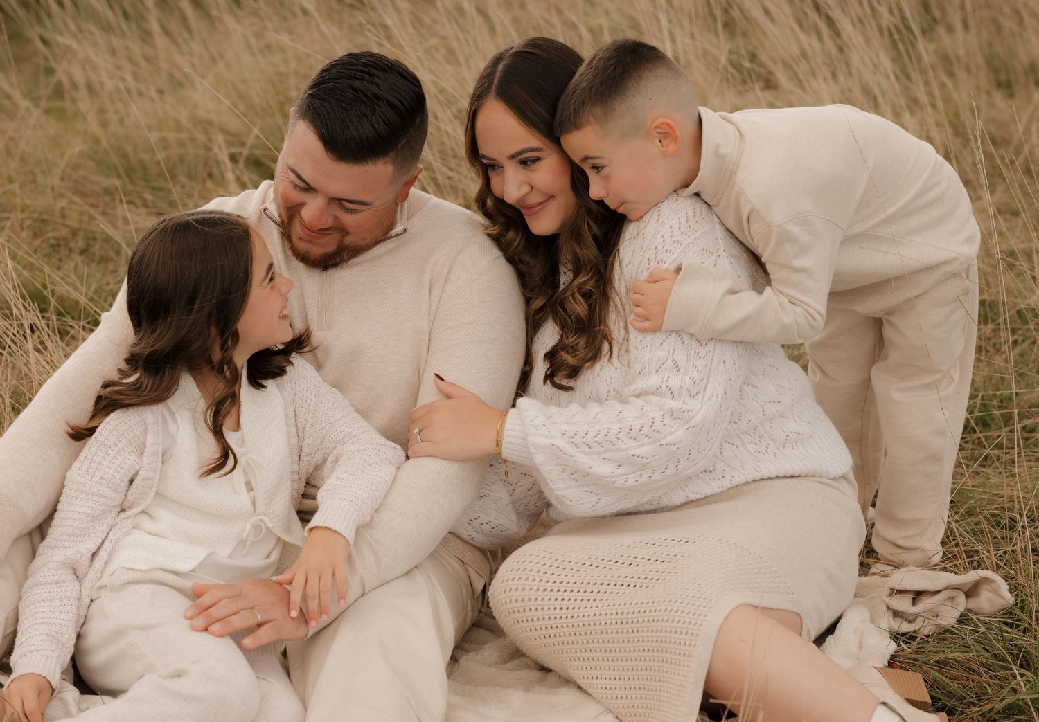 A family wearing coordinated cream and white outfits sits together lovingly in a grassy field.
