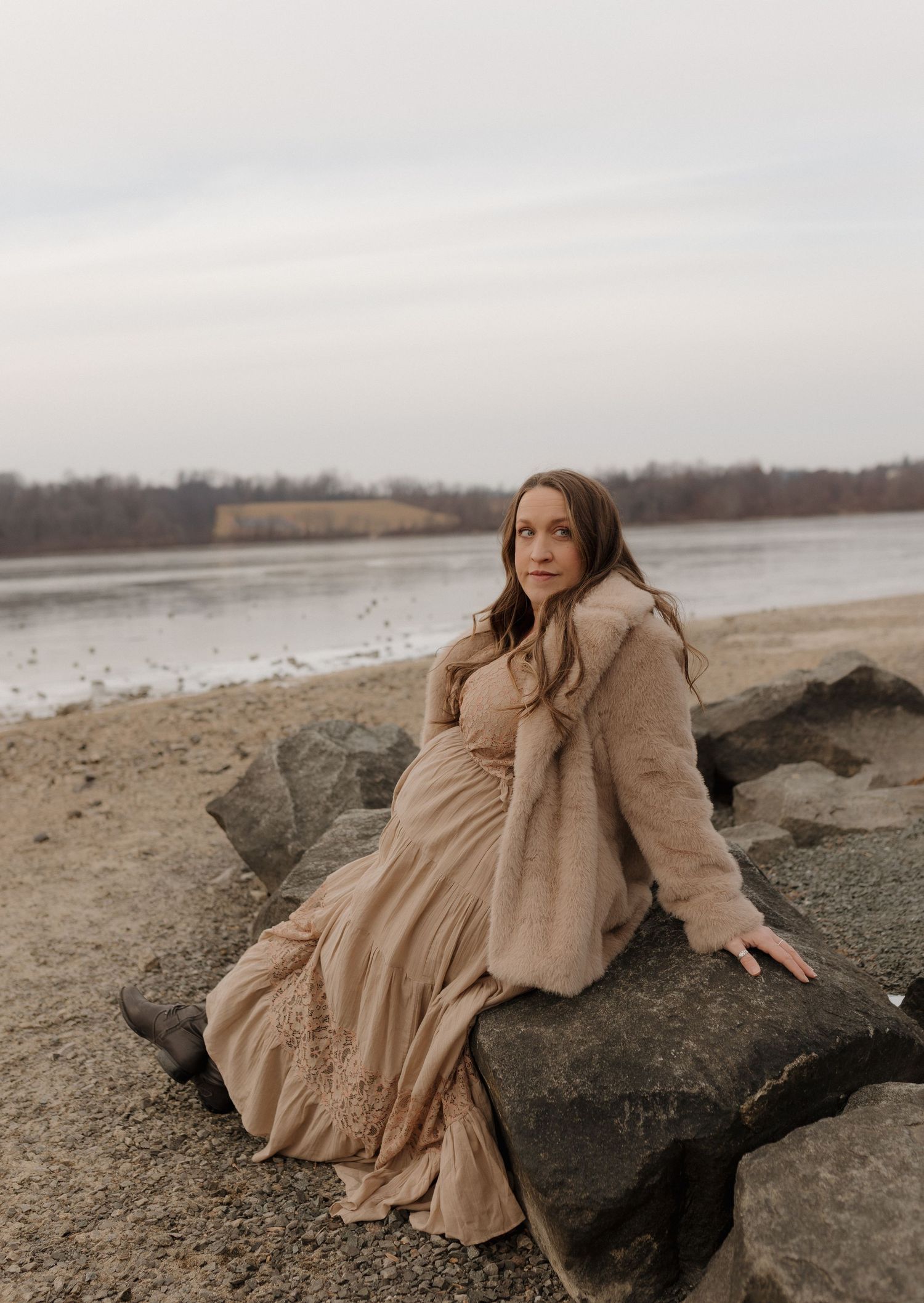 Woman in flowing beige dress poses dramatically on rocky shoreline during moody winter afternoon.