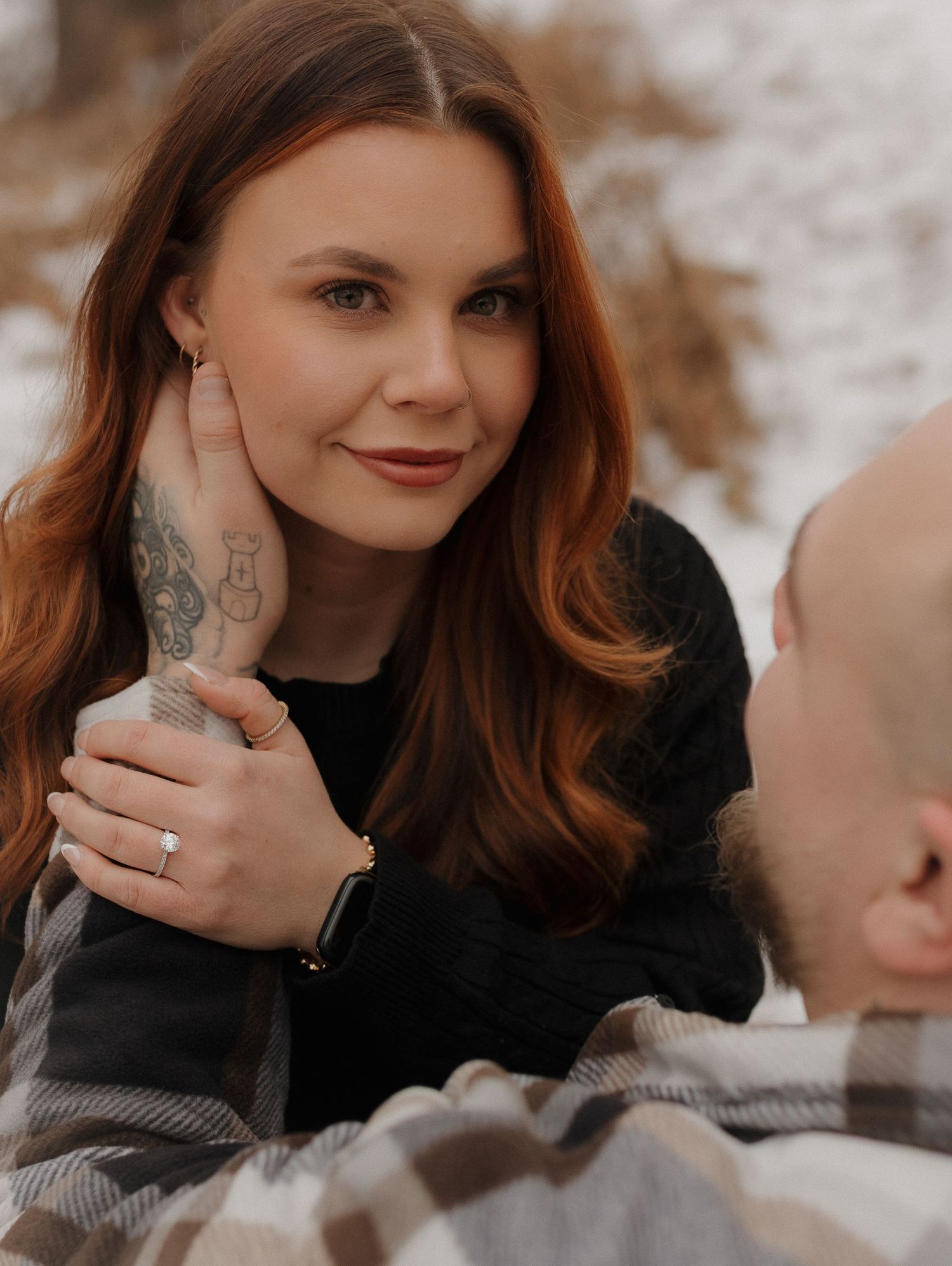 A woman with red hair wearing a black sweater poses outdoors with a visible tattoo on her hand.