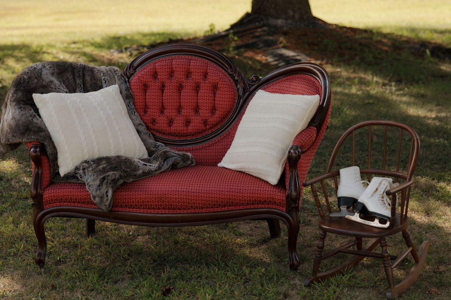 A red antique loveseat with white pillows arranged under a large tree.