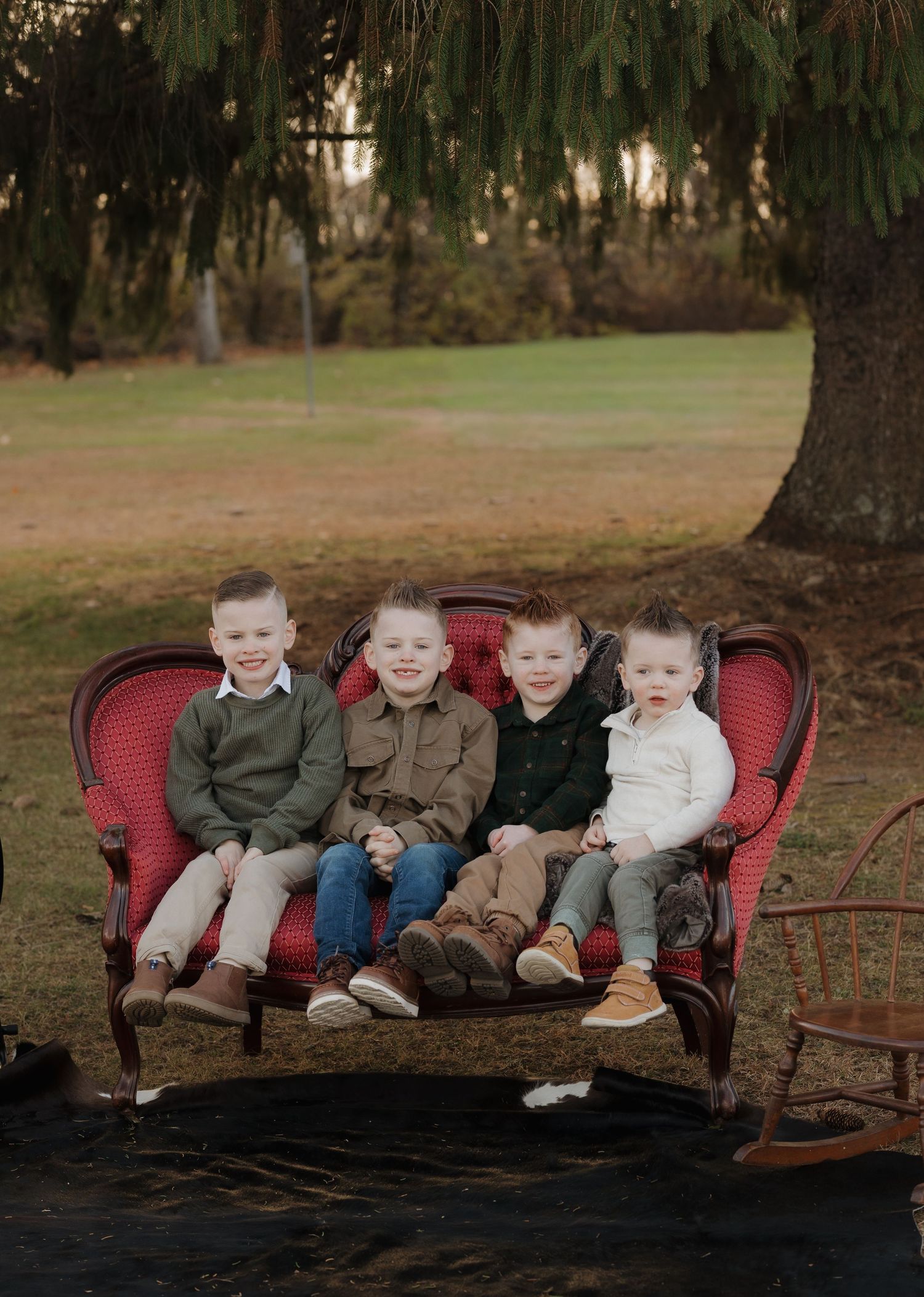 Four children sit together on a red vintage couch outdoors under a large tree.