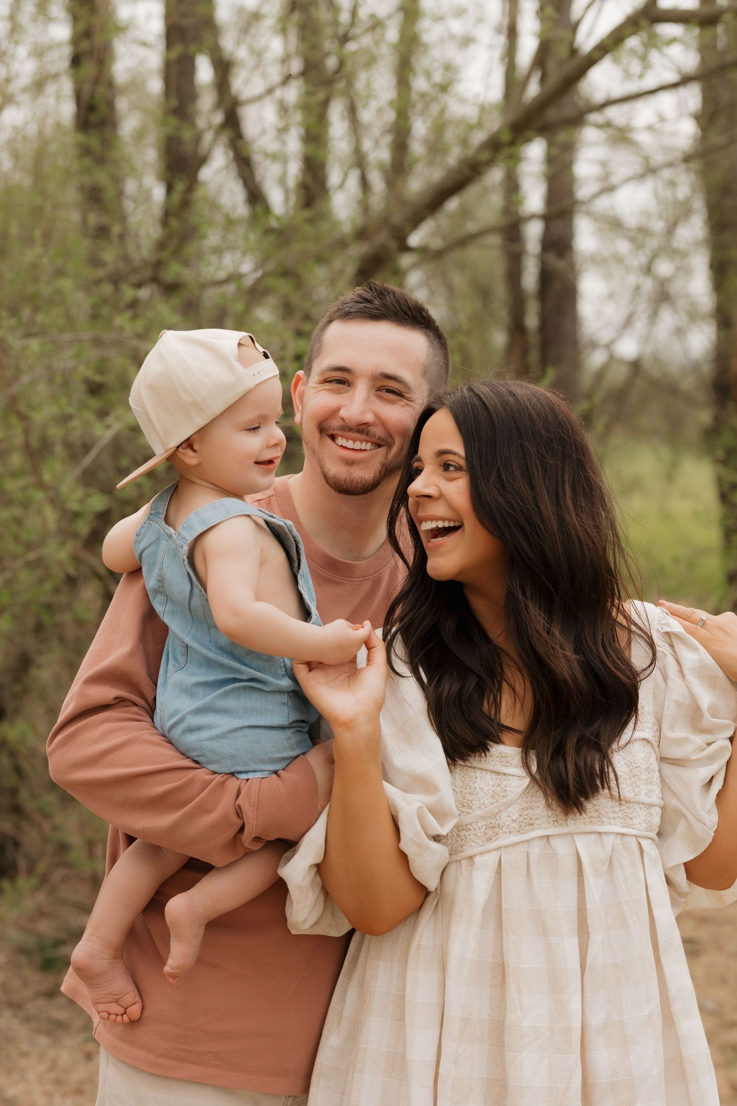 A family shares joyful moments together outdoors in a wooded setting wearing coordinated neutral-toned outfits.