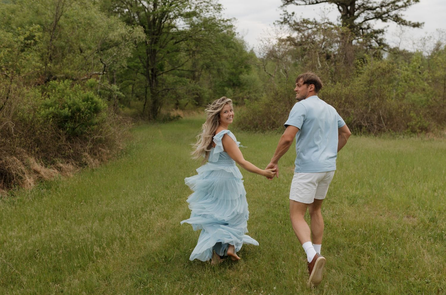 Couple holding hands running through green meadow, woman in flowing light blue dress and floral crown, man in blue shirt.