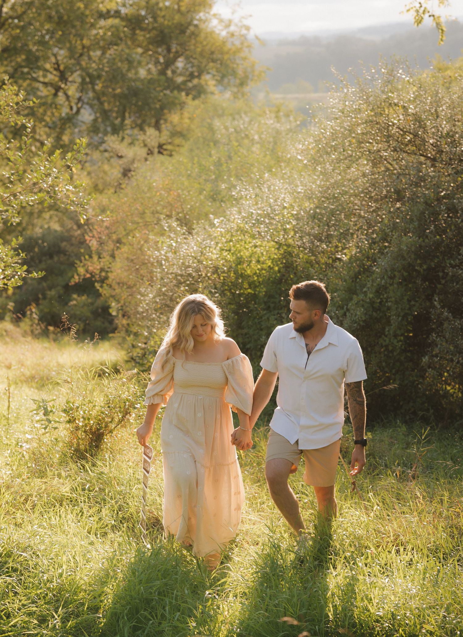 A couple walks hand in hand through a sunlit meadow at golden hour, surrounded by tall grass and natural foliage.