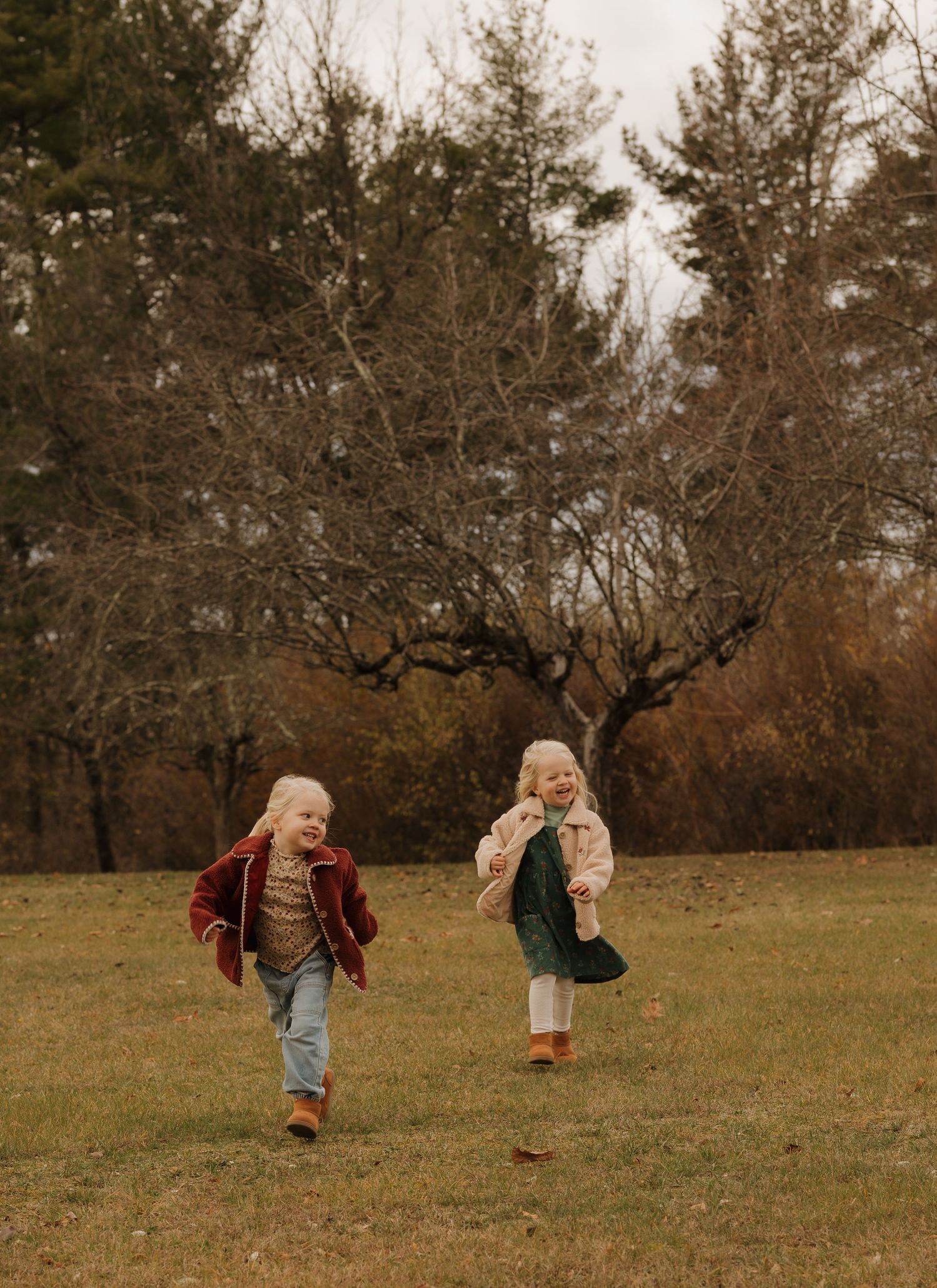 Two children run and play together in an autumn field surrounded by bare trees.