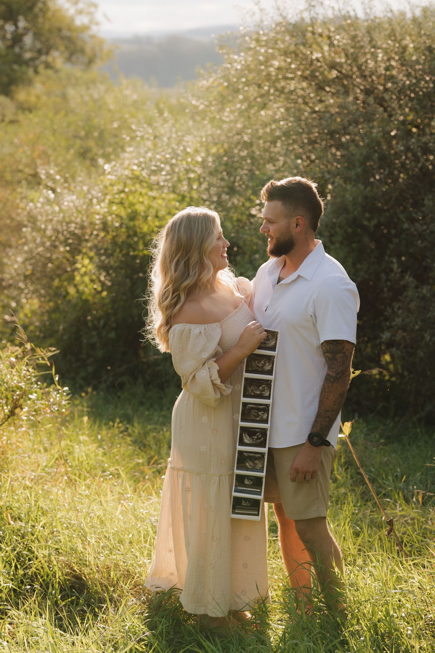 A couple shares a romantic moment in a sunlit meadow while holding ultrasound photos during their pregnancy announcement.
