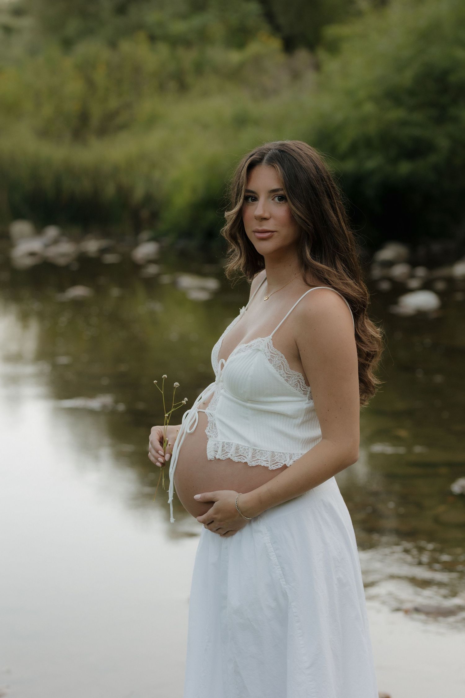 An expectant mother in a white dress poses by a serene lake.