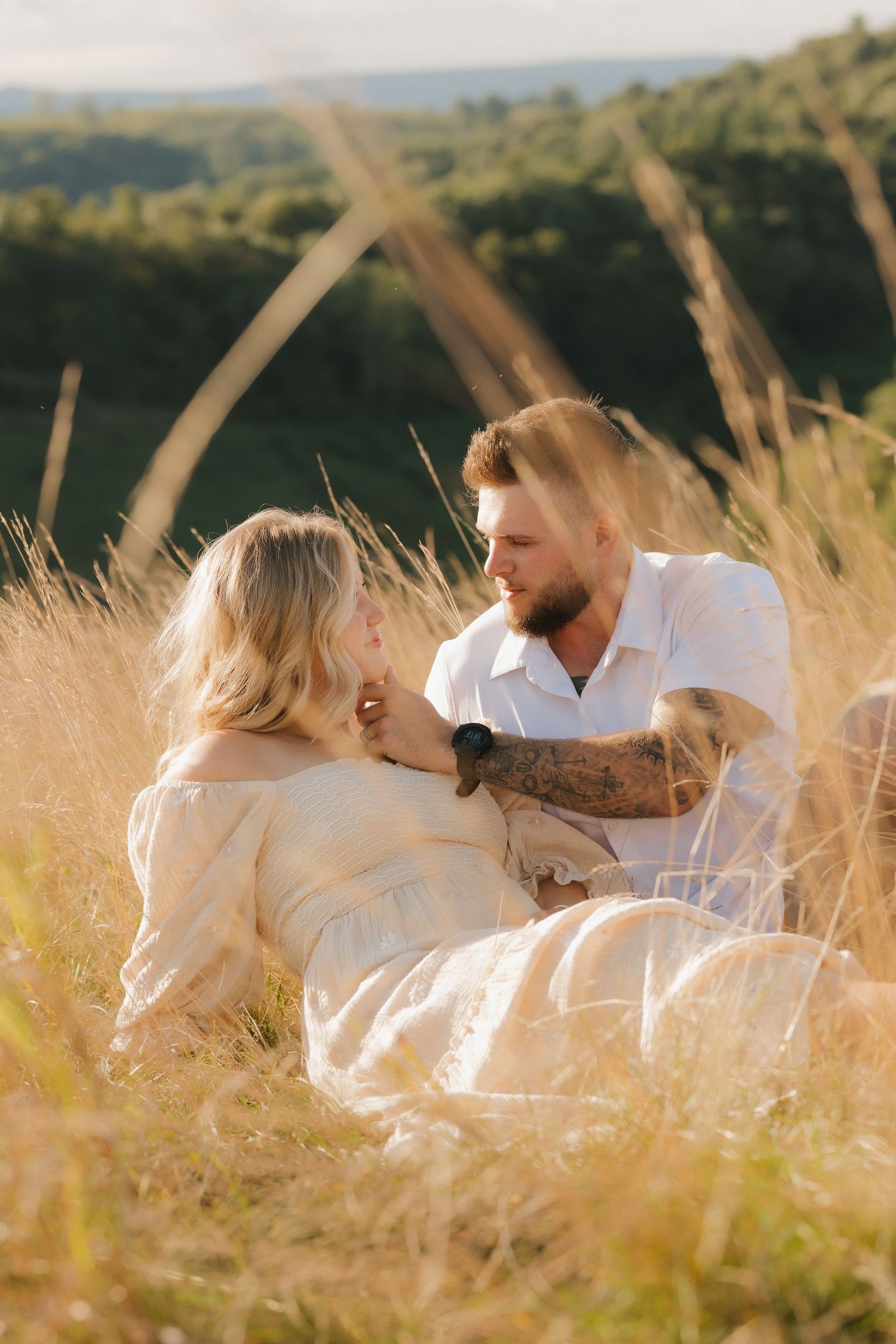 A couple in white outfits sits together in a golden wheat field during sunset creating a romantic summer scene.