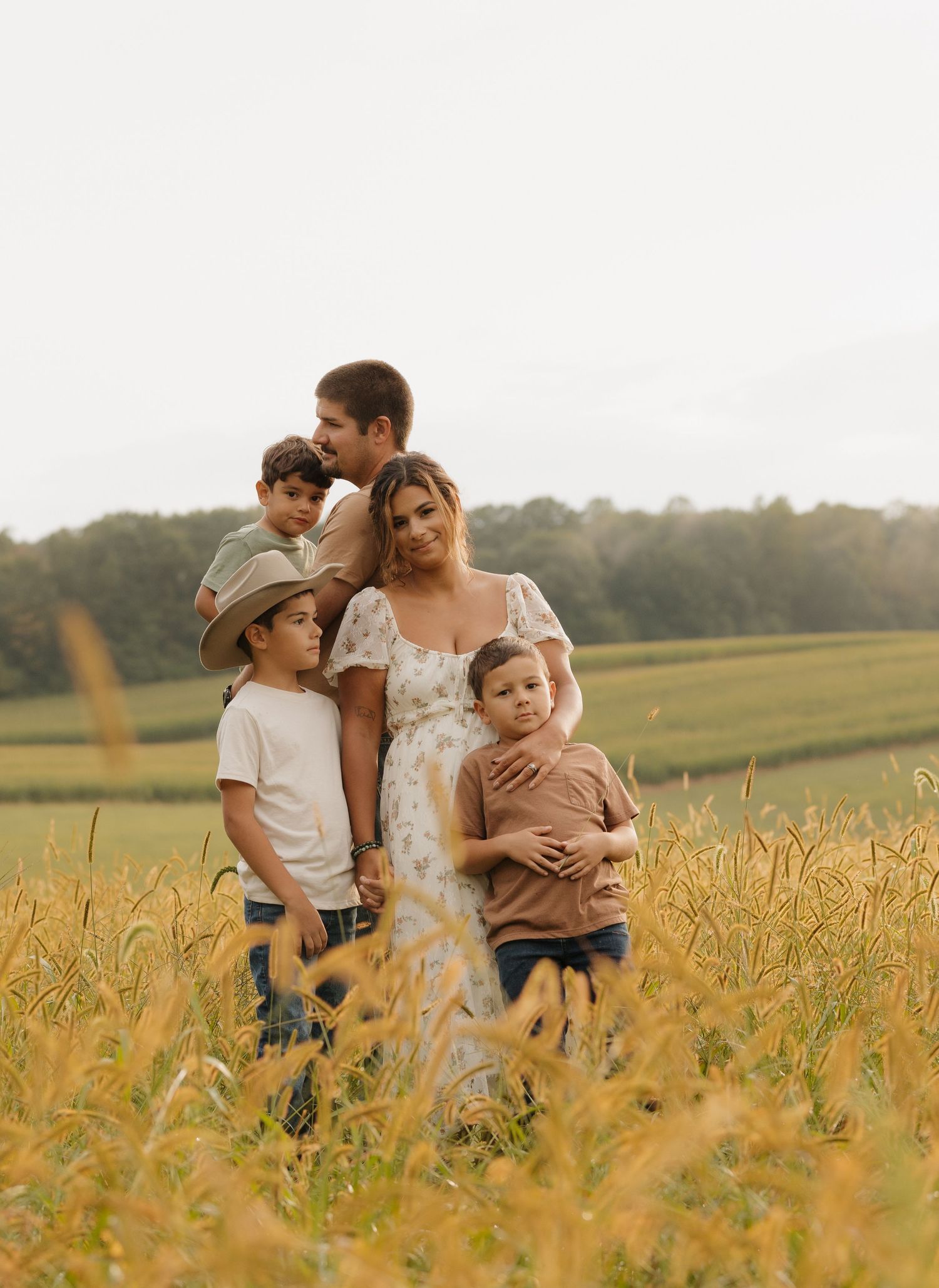 Family stands together in a golden wheat field at sunset with trees in the background.