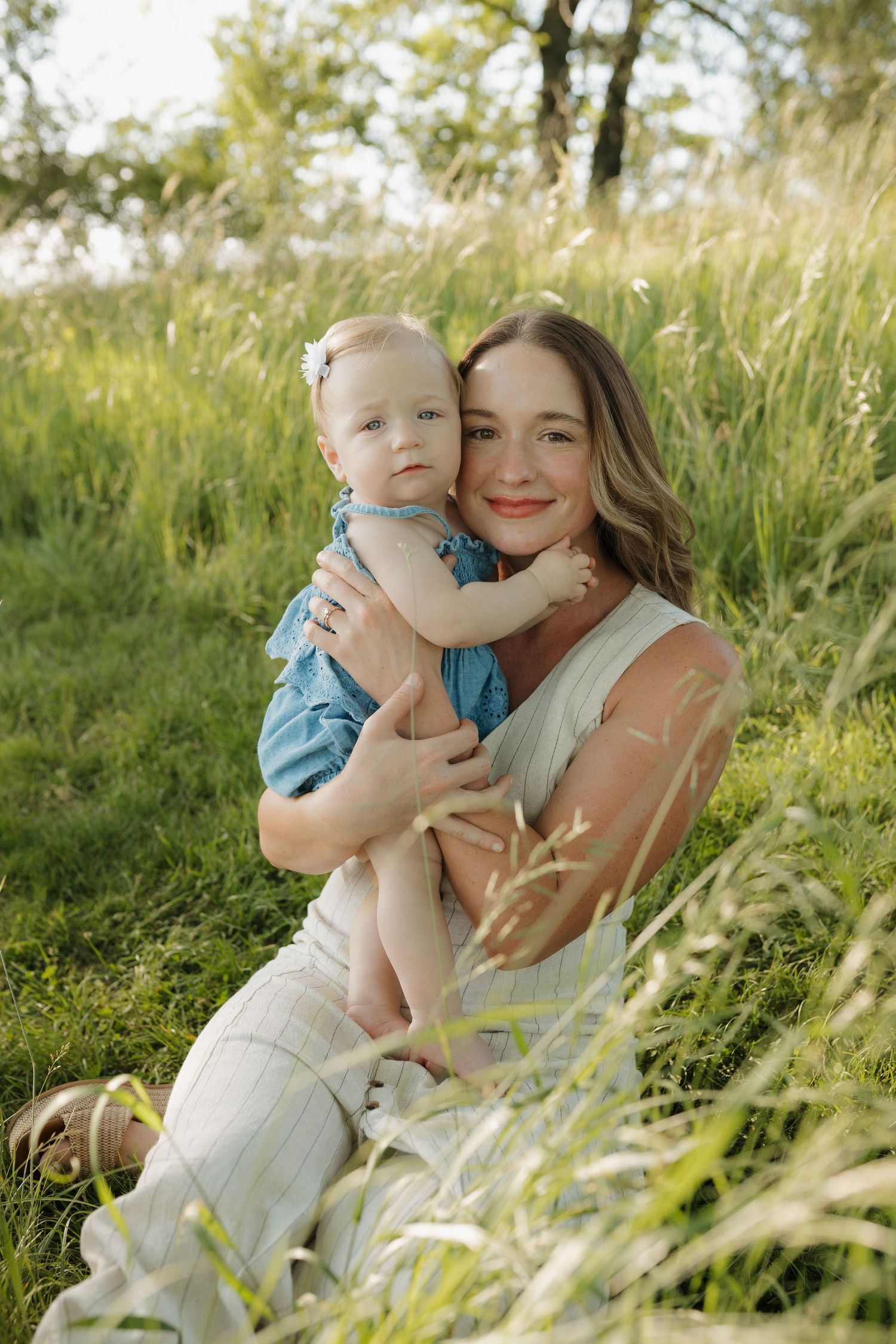 A smiling mother sits on green grass holding her baby girl in a blue romper during a sunny outdoor photo session.