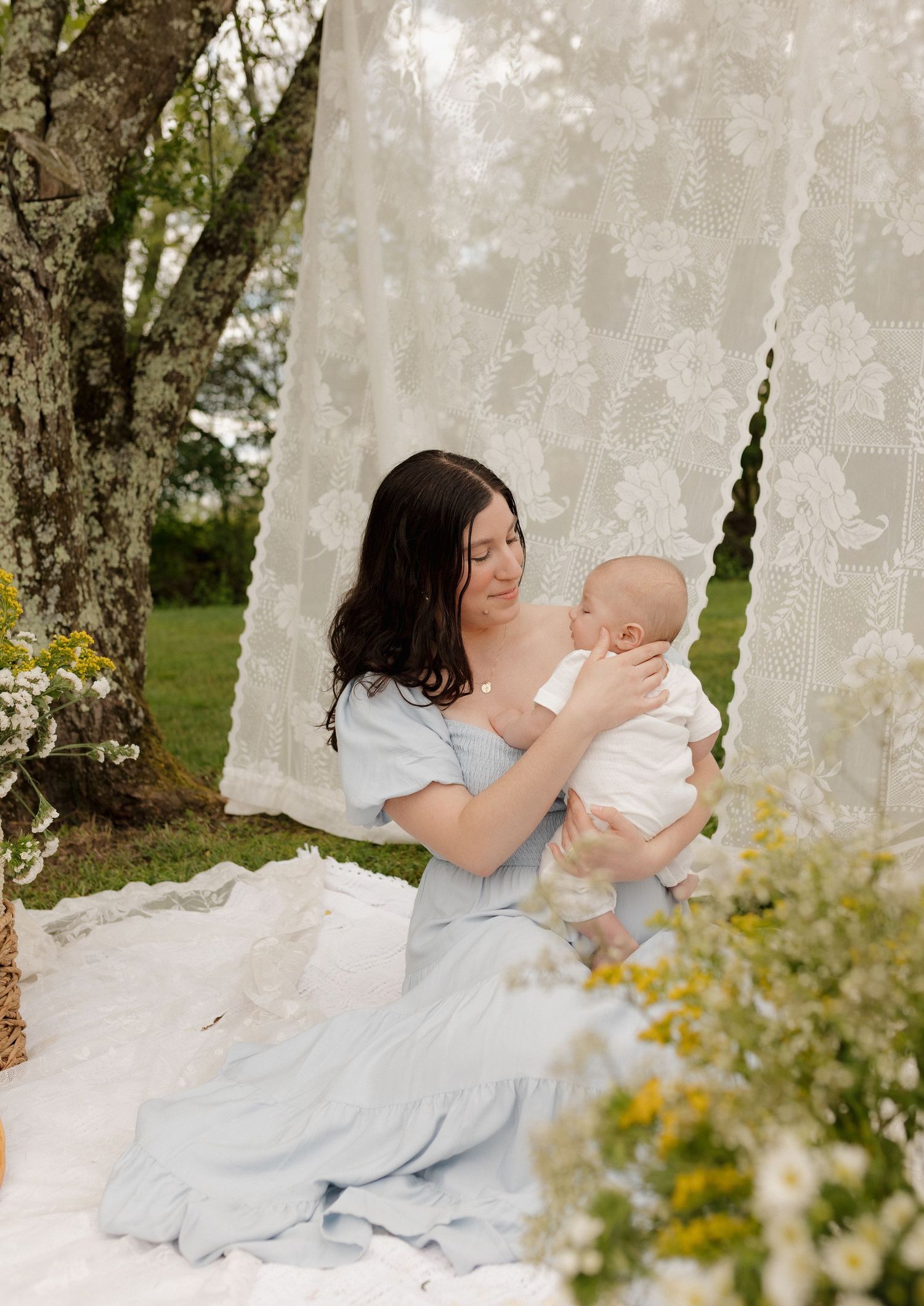 A mother in a blue dress holds her newborn baby outdoors under lace curtains surrounded by wildflowers.