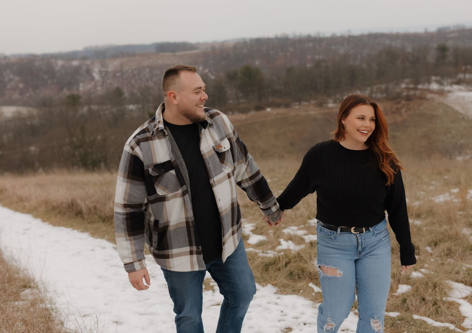 A couple walks hand in hand along a snowy path through a winter field with rolling hills in the background.