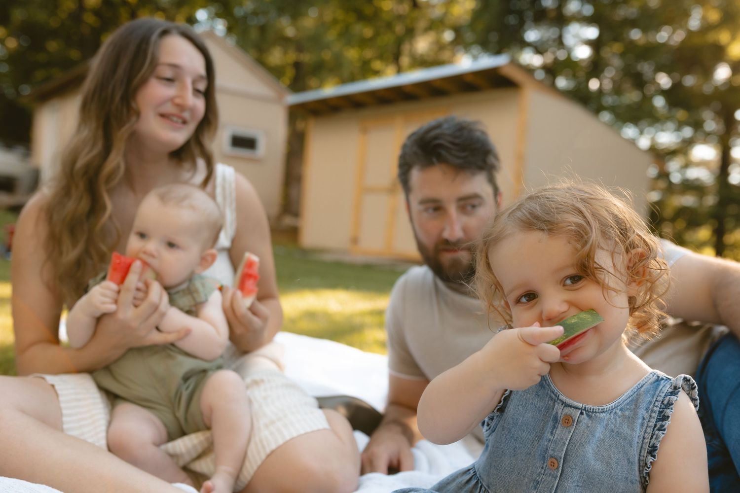 Family of four enjoying watermelon outdoors on a sunny day near a wooden shed.