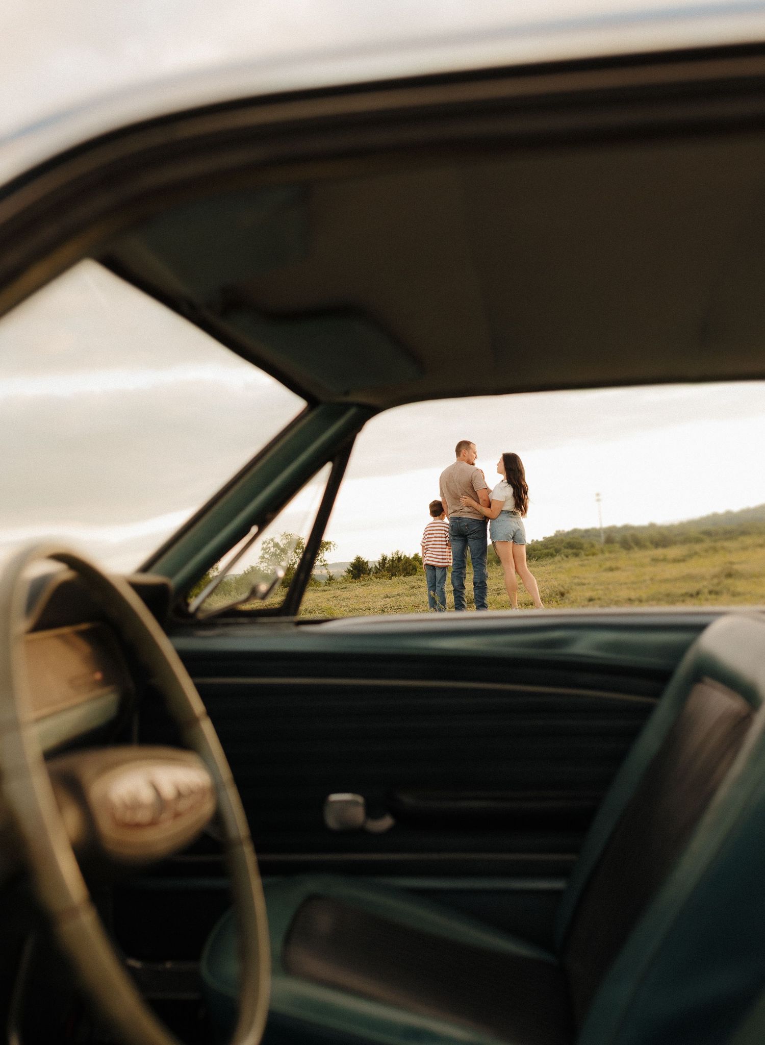 A family of three stands outdoors viewed through a vintage car window, framed by the classic steering wheel and dashboard.