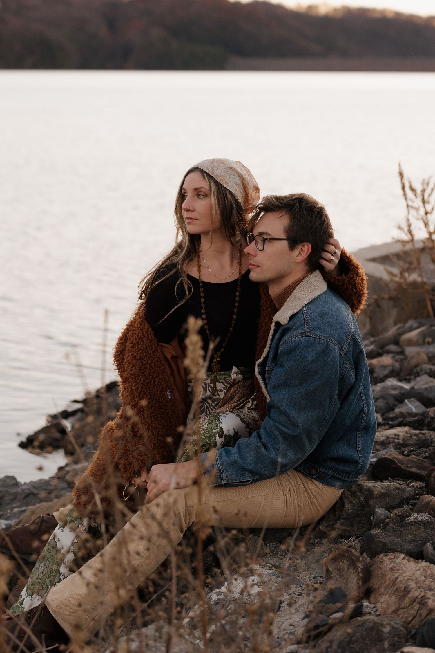 A couple sits together on rocky shoreline during sunset.