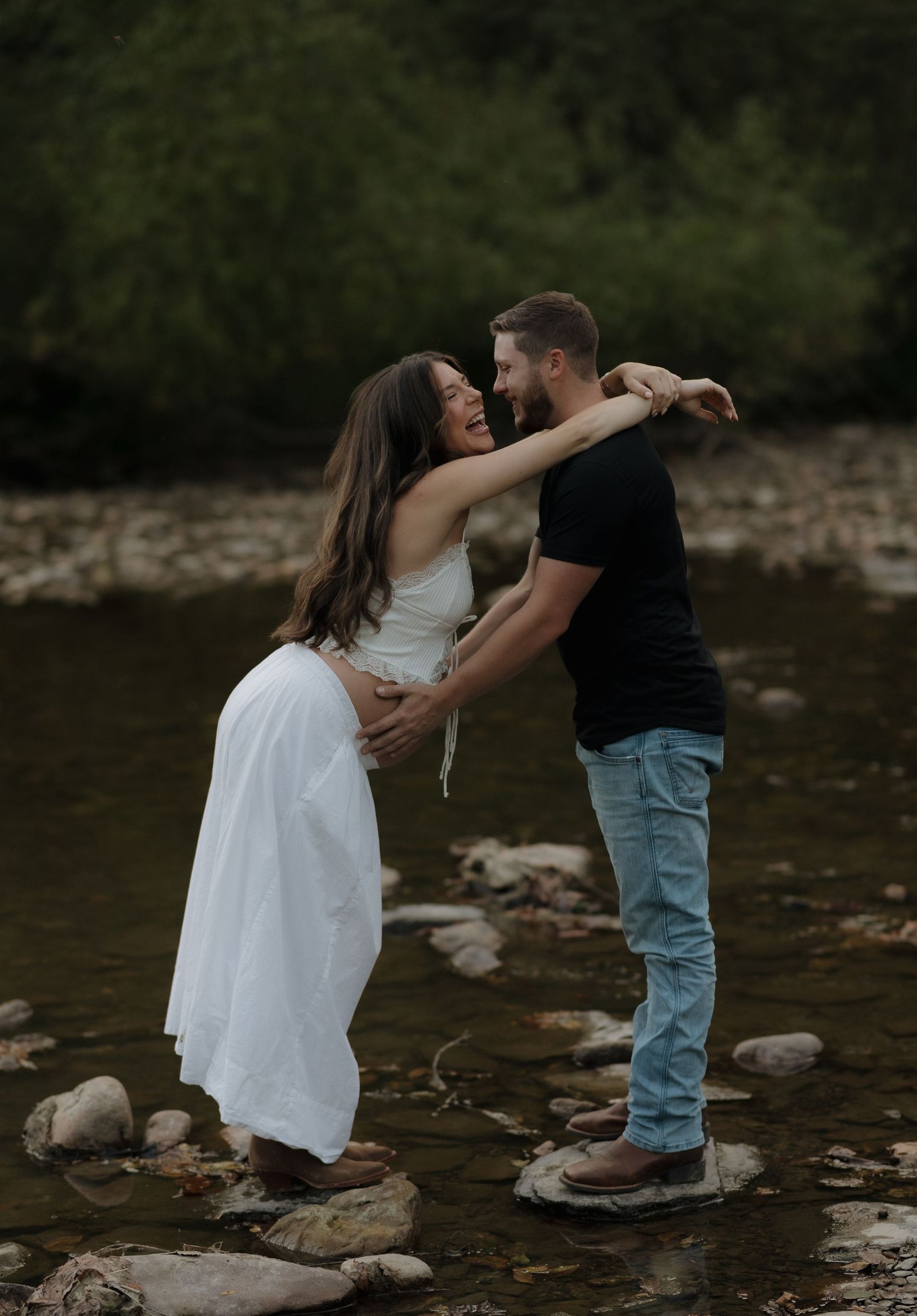 A couple embraces while standing on rocks in a stream during their maternity session.