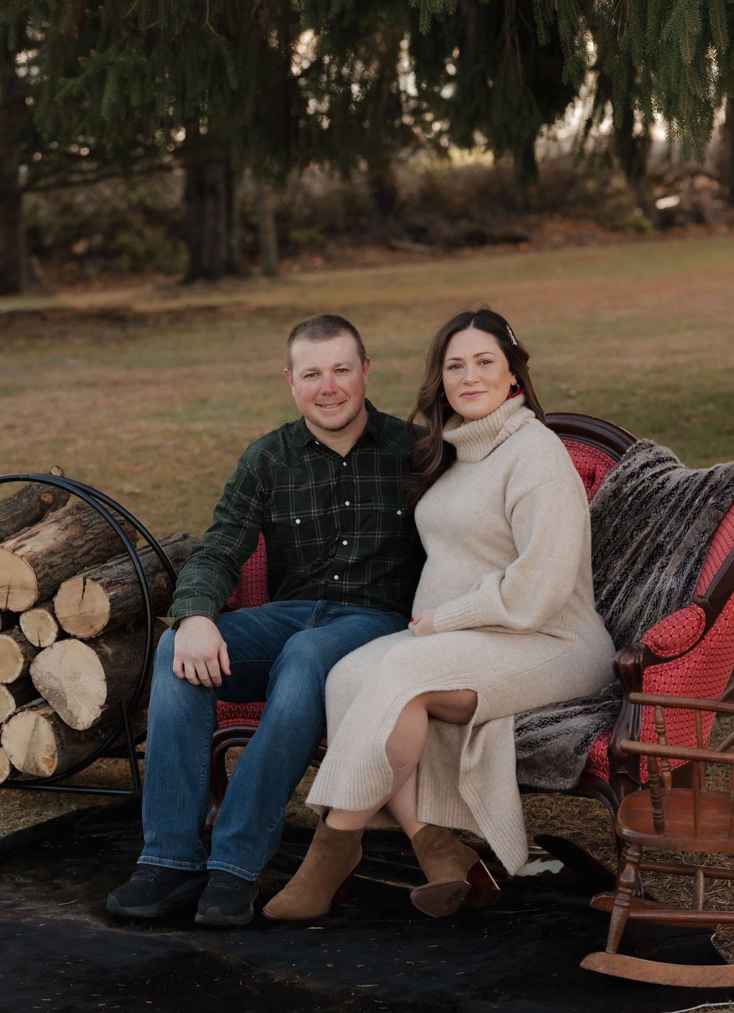 Couple sits together on a vintage red bench outdoors with firewood stacked nearby during a cozy photo session.